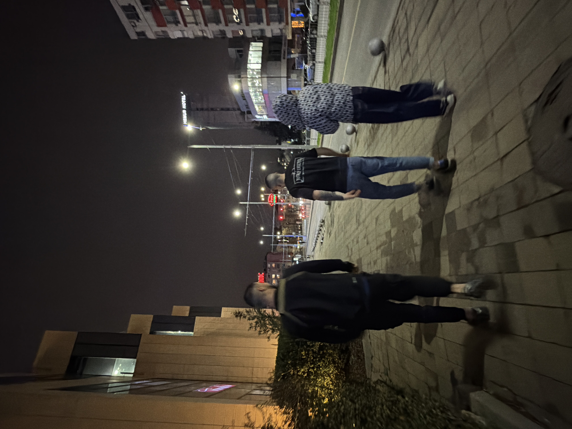 Three people are walking on a sidewalk next to a street at night in Sofia, Bulgaria. The Hemus Hotel sign is visible. There are balls on the sidewalk. Street lights and traffic lights are visible. There are buildings along the street.