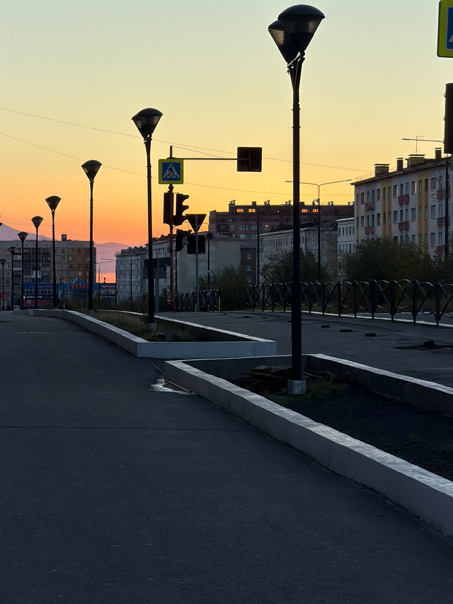 The image shows a street scene at dusk in Norilsk, Russia. The sky is a gradient of yellow and orange, indicative of a sunset or sunrise. Several streetlights with conical shades line the left side of the road, with one in the foreground being most prominent. A pedestrian crossing sign and a traffic light are visible on the right side of the road. In the background, multi-story apartment buildings with distinctive architectural features, common in Norilsk, line the street. Some of the buildings have illuminated windows. A low, decorative metal fence runs along a raised area on the right. The road surface in the foreground appears to be asphalt. The overall scene suggests an urban environment in Norilsk during a time of low light.