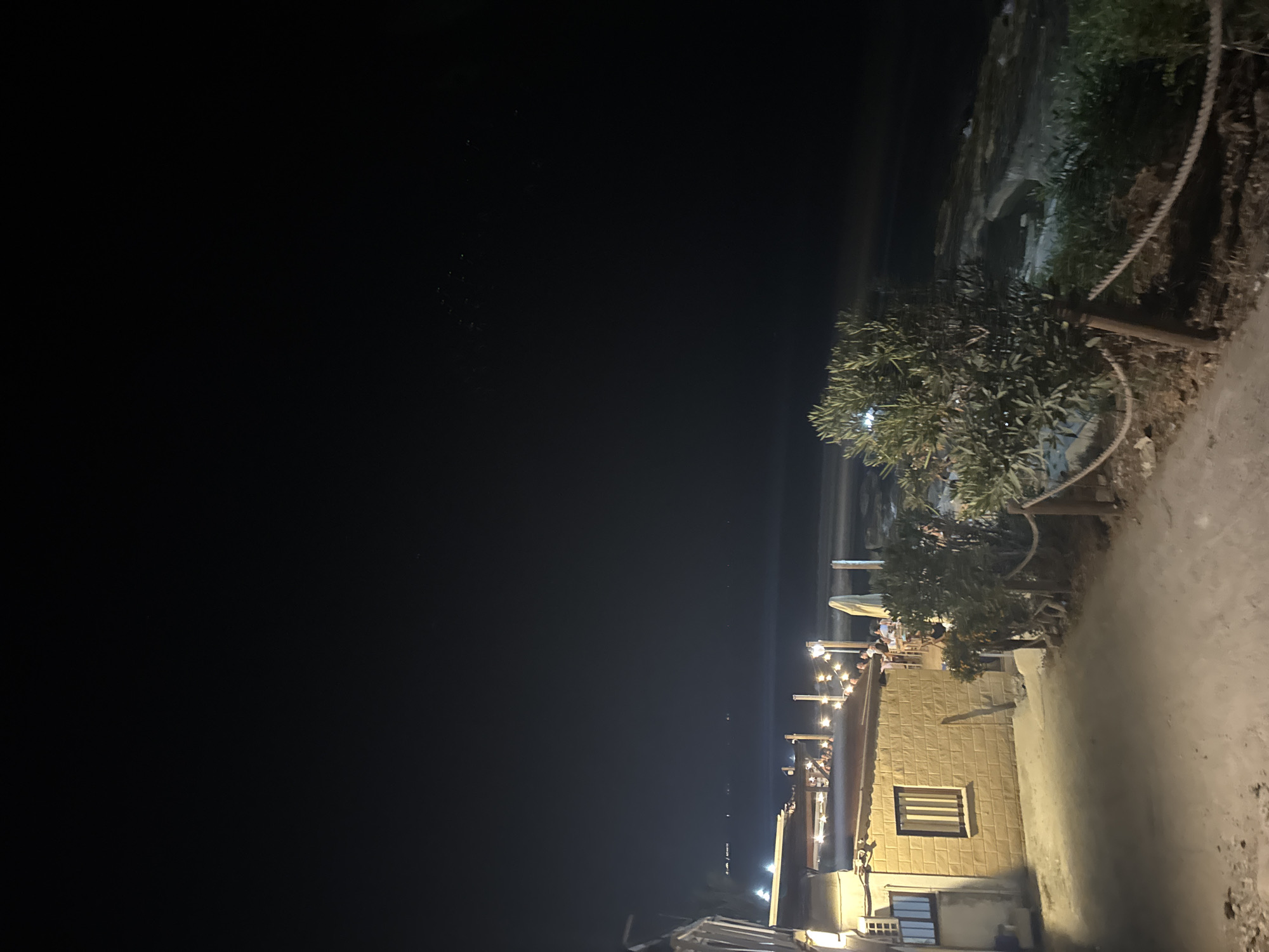 This image depicts an outdoor dining area at night in Agios Tychona Community, Cyprus. The scene is illuminated by string lights hanging over tables where several patrons are seated. To the left, a small building with a tiled roof and a window is visible, constructed with light-colored bricks. Adjacent to the building is a pathway leading towards the dining area and the sea. The sea is visible in the background, with some lights possibly from boats or distant structures visible on the horizon. A tree with green foliage is situated to the right of the pathway, with a rope barrier in front of it. The overall atmosphere suggests a restaurant or bar with a view of the sea.