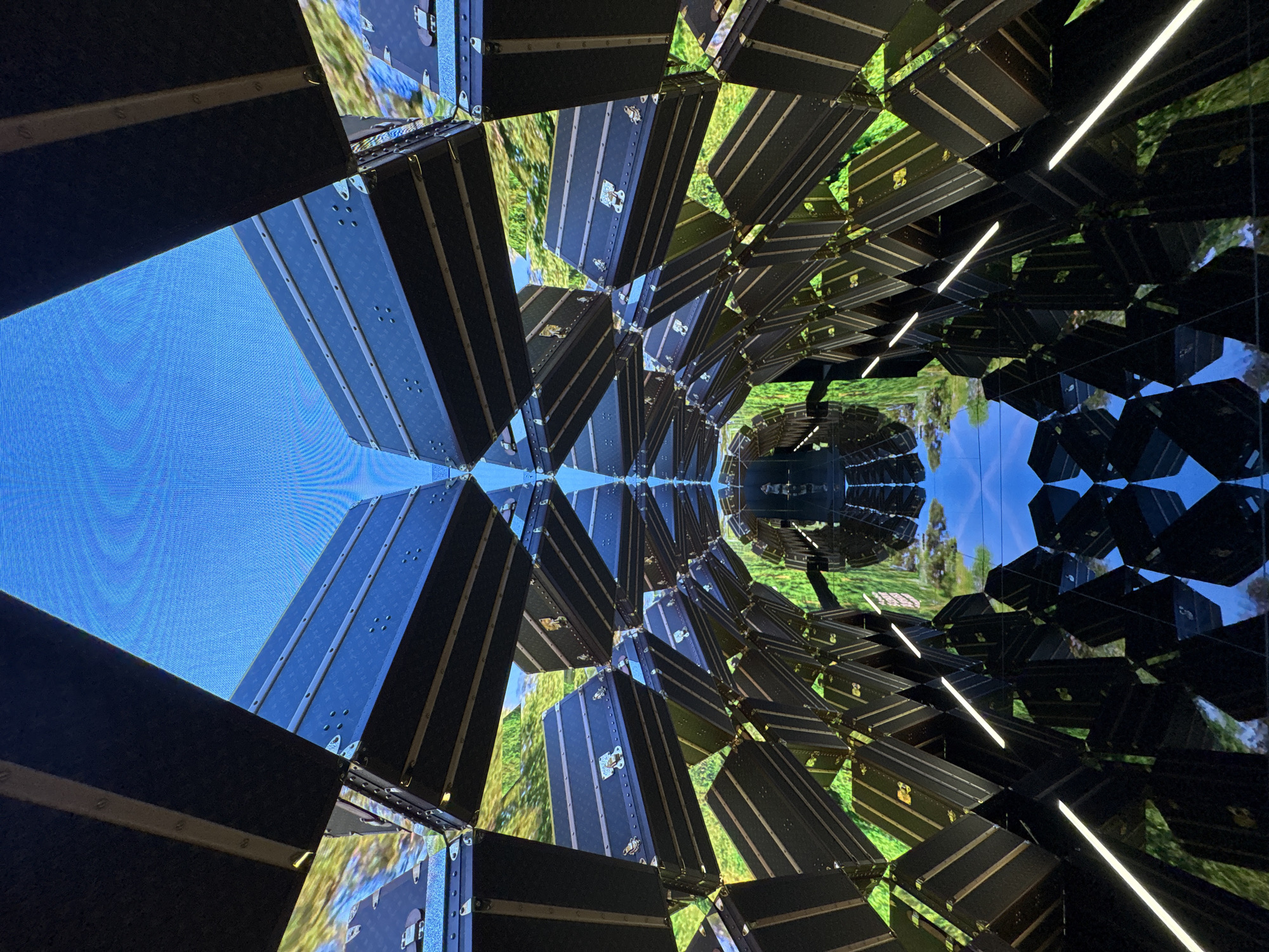 The image shows an abstract art installation, possibly in Bangkok, Thailand, featuring a series of vintage-style black trunks with metal accents, arranged in a geometric pattern resembling a honeycomb or kaleidoscope. The trunks are positioned at various angles, creating a visually complex and immersive environment. Some trunks appear to be mirrored or reflective, contributing to the kaleidoscopic effect. A blue screen backdrop is visible in some areas, suggesting an intentional design element. LED strip lights run along some of the geometric lines, enhancing the lighting and highlighting the structure of the installation. The reflections and the overall composition create a sense of depth and an optical illusion.
