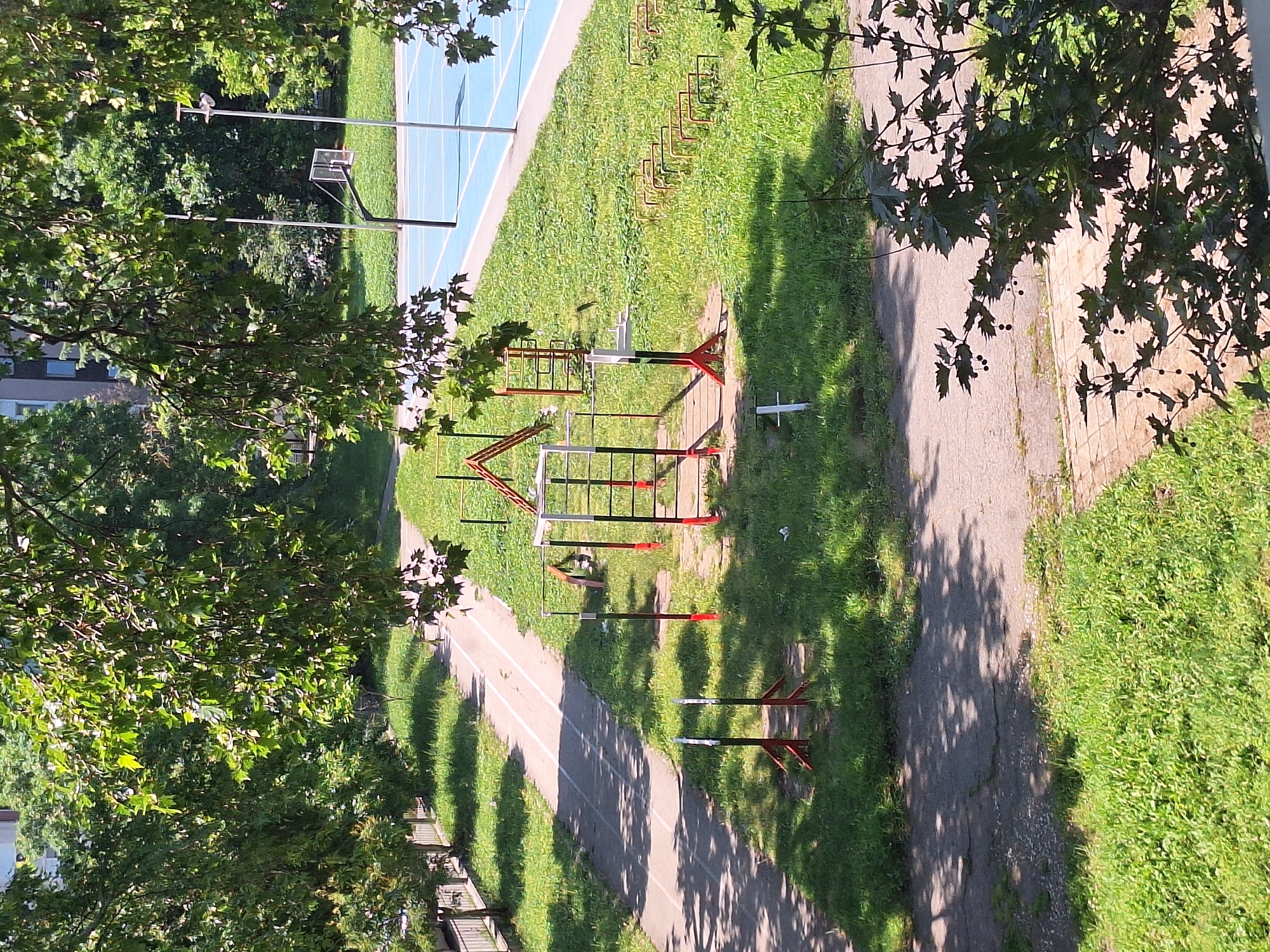 The image captures an outdoor sports and recreation area on a sunny day. In the foreground, through the dappled shade of tree branches, a paved pathway is visible, bordered by green grass. Further into the scene, a running track with white lane markings runs diagonally across the frame. Adjacent to the track is an open grassy area featuring outdoor exercise equipment. These include a multi-station calisthenics rig with red and white poles and horizontal bars, and a set of smaller red and white jump hurdles. Beyond this, a blue-surfaced sports court is visible, equipped with a basketball hoop and backboard. The background consists of lush green trees and a building. The overall impression is that of a public park or schoolyard. The image was taken in Varna, Bulgaria, in the vicinity of the Mladost residential area, specifically near the "Sv. Tsar Boris I" Secondary School.