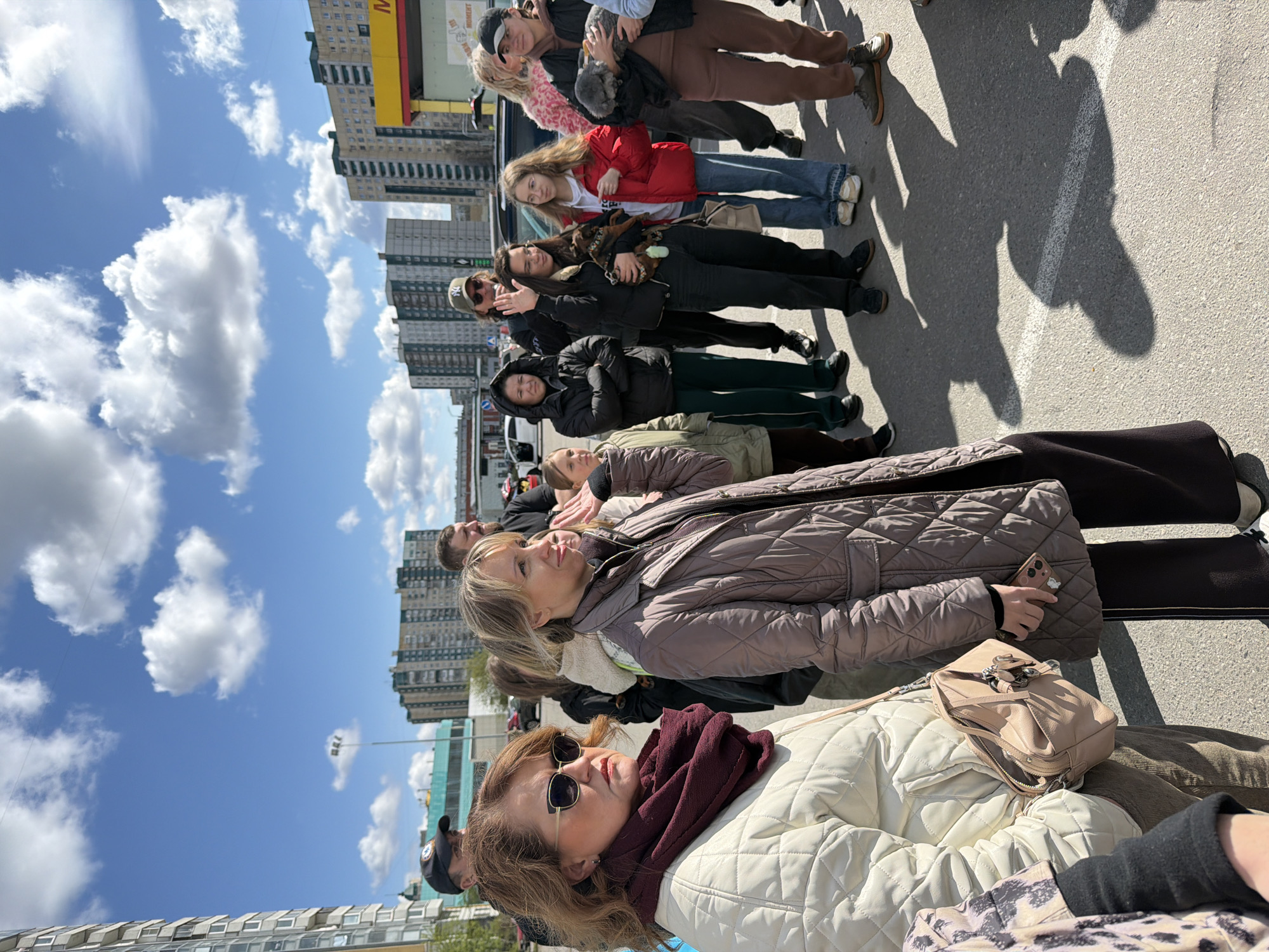 A diverse group of people, predominantly women and young adults, are depicted standing in a queue or line outdoors in an urban environment, likely in Saint Petersburg, Russia. The image captures a bright, sunny day with a clear blue sky adorned with scattered white cumulus clouds. Strong, defined shadows are cast on the asphalt ground, indicating midday sun.
Individuals in the line are dressed in casual outerwear, suitable for cool to mild weather, including various quilted jackets in shades of brown, cream, and black, as well as parkas and coats. Accessories such as sunglasses, smartphones, handbags (including a beige quilted one and a small tan shoulder bag), and baseball caps are visible. One person is seen cradling a small pet, possibly a dog, wrapped in a blanket, while another holds a fluffy, grey object. The expressions on their faces are mostly neutral, suggesting patience as they wait.
In the background, several tall, multi-story residential or commercial buildings rise, providing a typical urban backdrop. One building features a partially visible yellow and red sign with a prominent Cyrillic letter "М", likely indicating a commercial establishment common in Russia. The scene conveys an everyday urban gathering or waiting experience under clear skies.