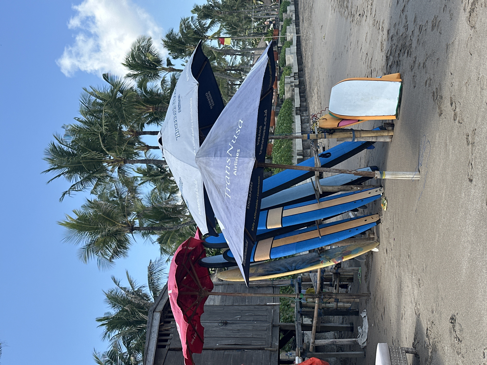 This is a medium shot of a beach scene in Kuta, Indonesia, dominated by surfboards and umbrellas. In the foreground, several surfboards are propped upright and lean against a wooden structure. The surfboards display a variety of colors and patterns, with prominent blue and yellow sections. Two large umbrellas are open, providing shade. One umbrella is a light blue color with "TransNusa Airlines" printed on it, along with "SINGAPORE" and "Safe Affordable Comfortable". The other umbrella is dark blue, also branded with "TransNusa Airlines" and text including "MANADO SORONG". A red umbrella is partially visible to the left.
Behind the surfboards and umbrellas, a rustic wooden structure, possibly a beach hut or lifeguard station, is visible. Palm trees are abundant in the background, reaching towards a clear blue sky with a few wispy clouds. A small flag with red and yellow sections is planted in the sand further back. The beach itself is sandy, with footprints visible in the foreground. In the far left of the frame, a piece of outdoor furniture, possibly a lounge chair, is partially visible. The overall impression is a commercial beach setup in a tropical location.