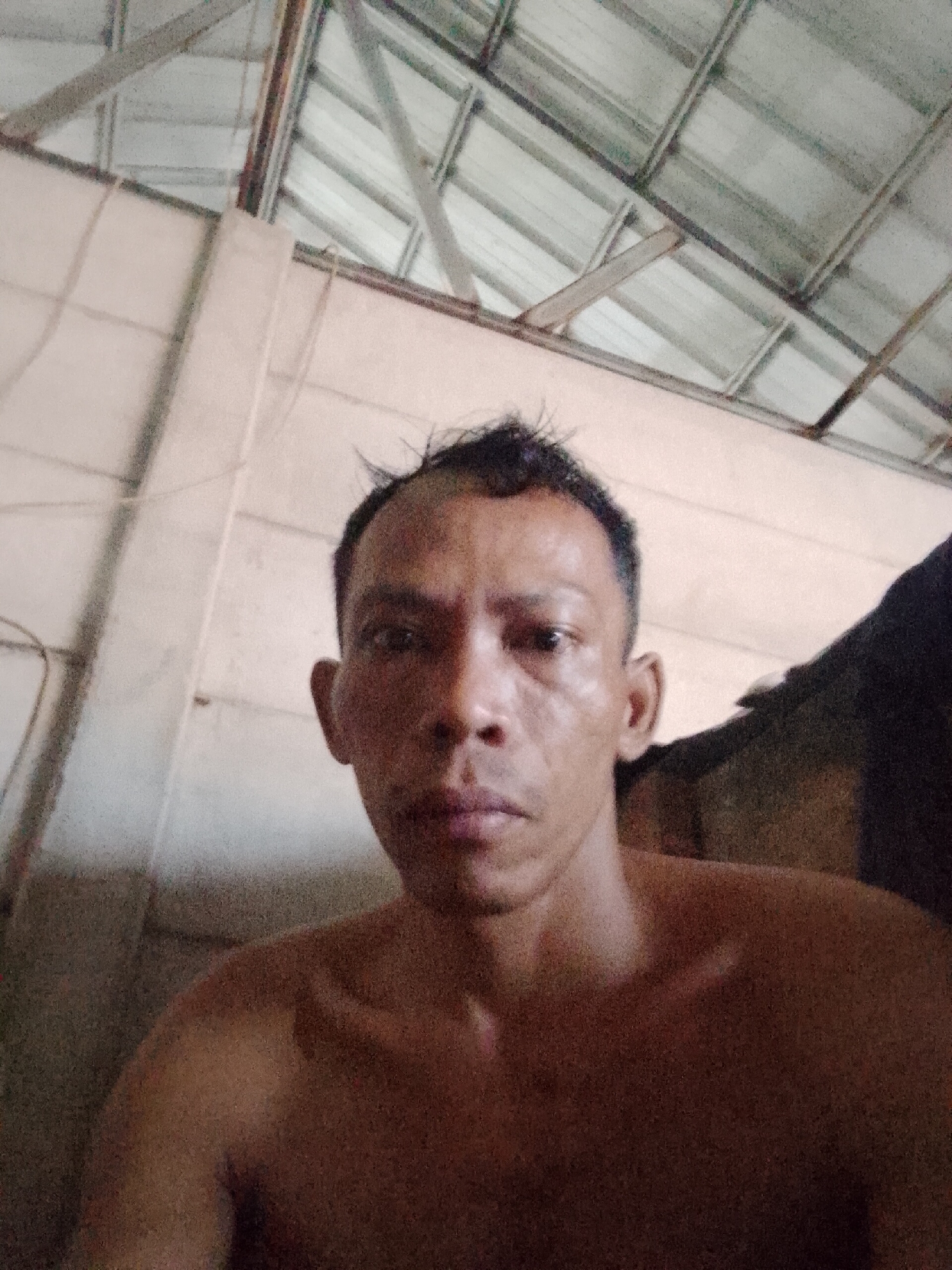 A medium, eye-level indoor selfie of an adult Asian male with dark skin and short black hair. He appears shirtless. The background consists of a white wall on the left and center. A metal ceiling structure is visible above. The location is in Bukit Ketok, Indonesia.