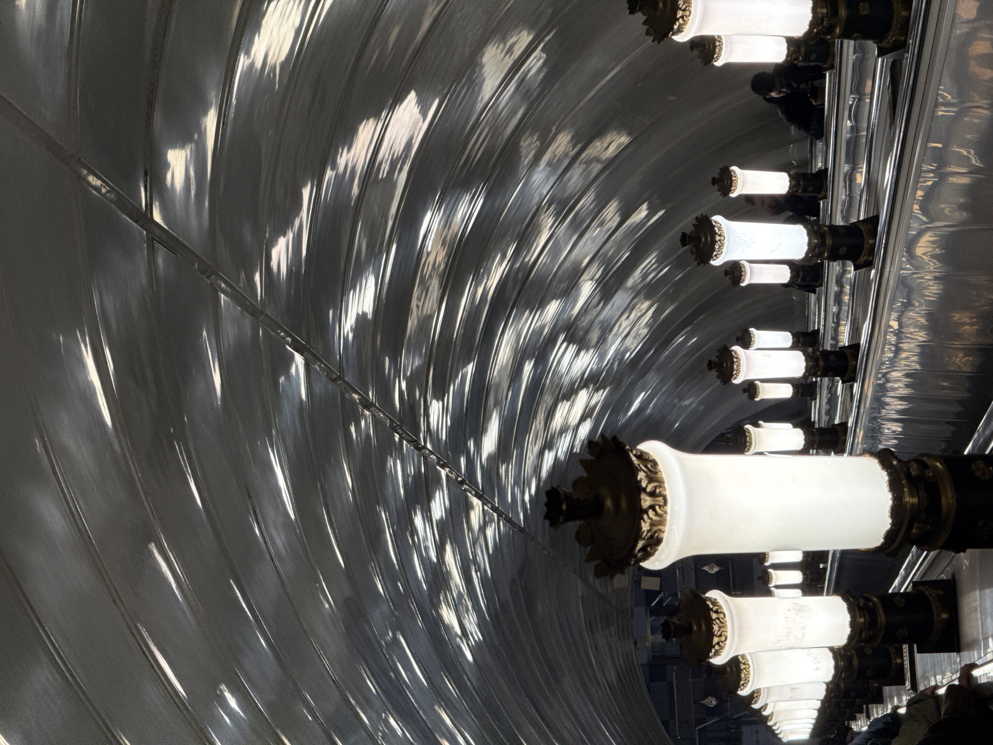 The image captures an interior space, likely a metro station in Moscow, Russia, characterized by a curved, reflective ceiling with a ridged texture that amplifies the play of light. A row of decorative, cylindrical light fixtures lines the right side of the frame. The light fixtures have metallic bases, and cast a warm glow. Several people are visible on the right, and they are in the lower section of the image. The composition emphasizes the architectural elements and the interplay of light and shadow within the space.