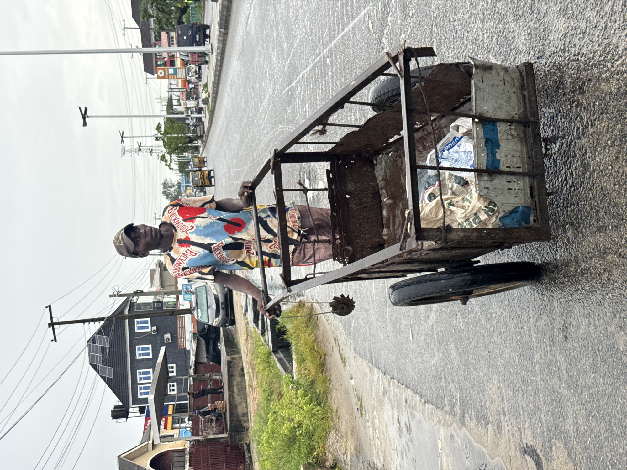 A man wearing a patterned shirt and cap is pulling a rusted, metal cart filled with items, including what appears to be bags of trash, on a street in Ado, Nigeria. The street is paved, slightly wet, and lined with buildings, power lines, and parked vehicles. A drainage canal with overgrown vegetation runs alongside the road near the man. The sky is overcast.