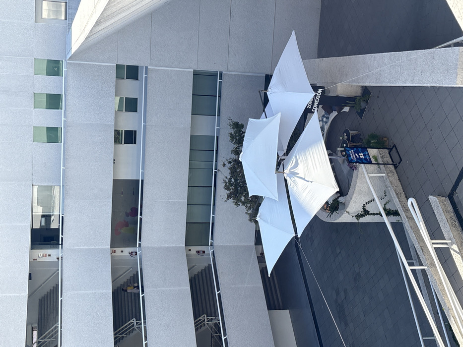 The image depicts an outdoor courtyard or patio area situated within a modern, multi-story building complex in Heroica Puebla de Zaragoza, Mexico. The architecture features light grey, textured concrete facades with prominent open-air walkways, multi-level staircases, and numerous windows.
In the paved courtyard at the lower level, four large, white, square patio umbrellas are open, casting shadows. These umbrellas are arranged around a curved, low stone wall that delineates an outdoor seating area. This area contains cushions, small circular tables, and potted plants. A black sign with white text reads "ZONA CONEXION" and "LIVE THE EXPERIENCE," identifying the space. Another smaller black sign with blue and white graphics is also present. Green vines grow on the curved stone wall, and a tree with green foliage is visible behind the umbrellas near the building.
On the upper levels of the building, which include multiple balconies and walkways with white railings, specific details are discernible. On one of the intermediate open-air levels, a female individual, dressed in a light-colored top and dark pants or skirt, is seen ascending an outdoor staircase. On the same level, behind a large glass window or partition, two indistinct human figures are visible along with several red and yellow balloon-like objects or decorations. At least two red fire extinguishers are mounted on walls adjacent to the staircases on different levels of the building. The building incorporates a variety of window types, including reflective, tinted green, and clear panes. An angular, upward-sloping concrete architectural element is visible on the upper right side of the structure.