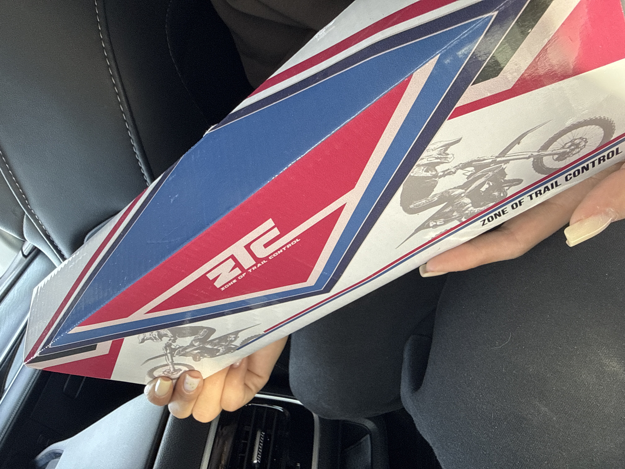 The image shows a person sitting in the driver's seat of a car, holding a box labeled "ZTC Zone of Trail Control". The box has a red, white, and blue design with an image of a motorcyclist. The person is wearing black clothing. The car seat is black leather with white stitching. The car's interior features a black dashboard. The location is unclear, as it is an interior shot and Chaikovsky, Russia, cannot be determined from the visual information provided.