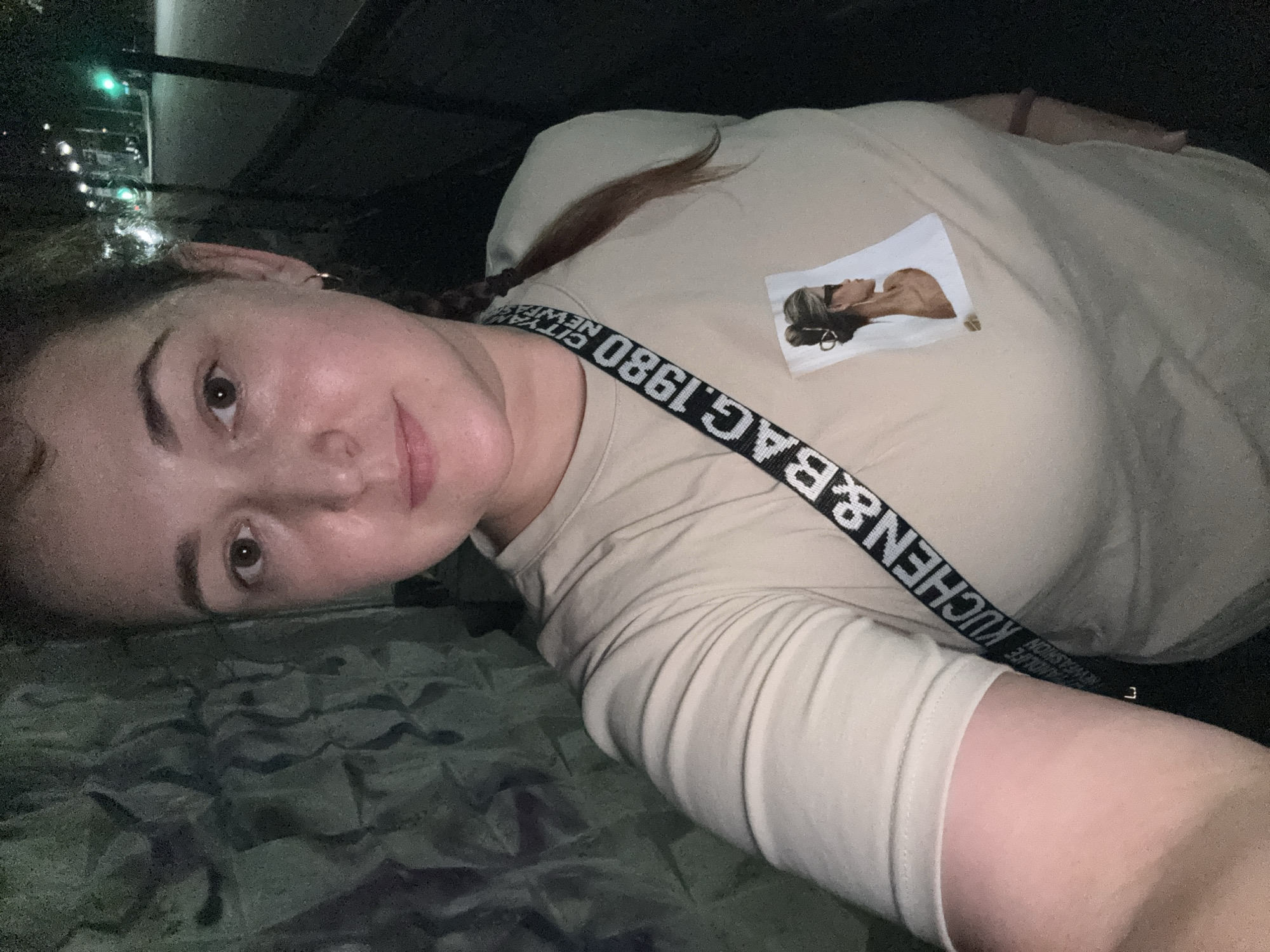A young woman with brown hair in a braid is taking a selfie at night. She is wearing a light-colored t-shirt with a picture of a woman on it and a black strap with white lettering across her chest. The strap reads "KUCHEN&BAG.1980 CITY NEWYORK FASHION." The background shows a dark, textured wall on the left and a street with lights and a traffic signal on the right. The location is identified as Chelyabinsk, Russia.