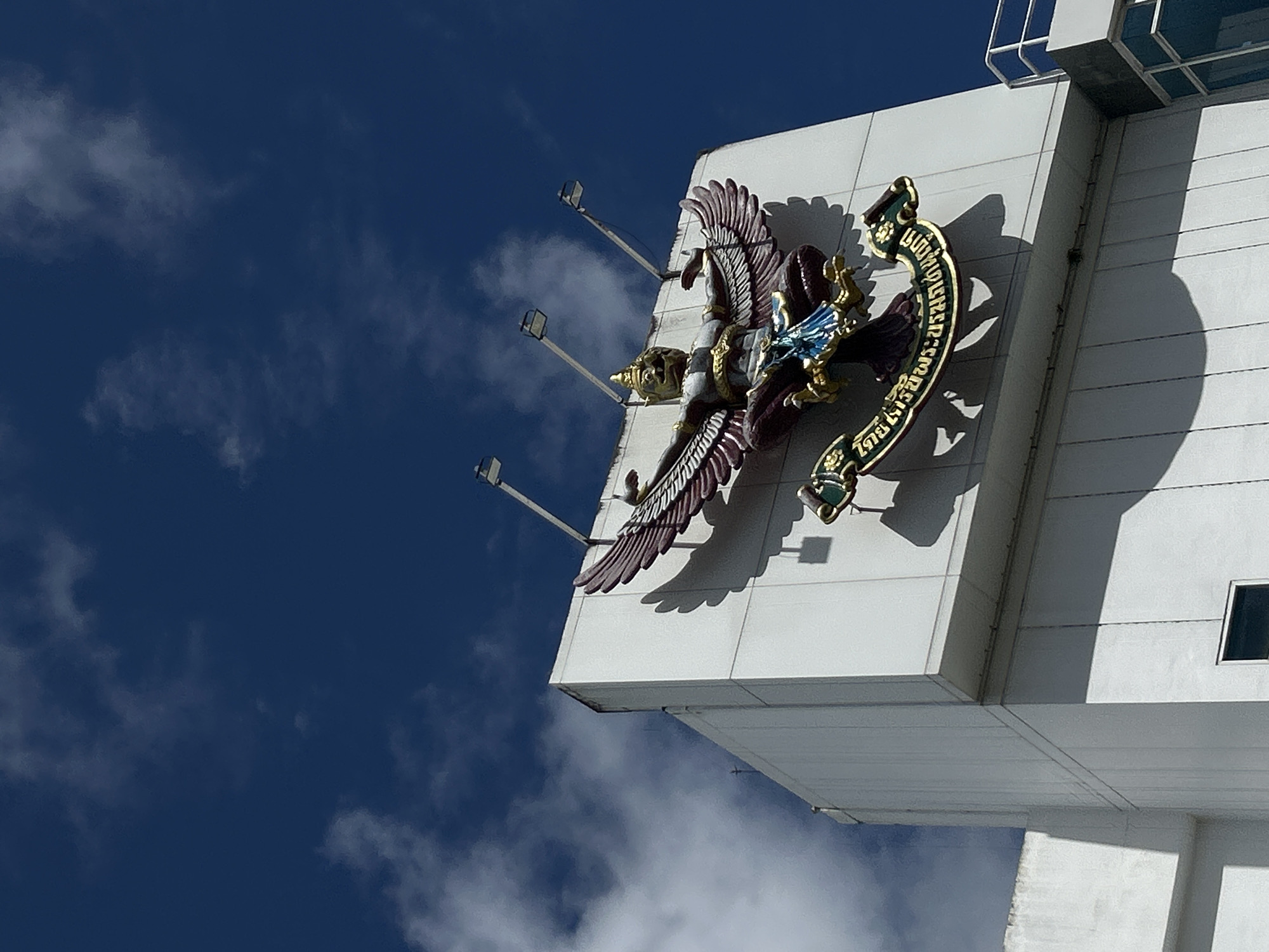 A statue of a Garuda, a mythical bird-like creature, is mounted on the exterior of a white, rectangular building. The statue is adorned with intricate gold and blue details, and its wings are spread wide. The building features several lights pointing upwards towards the sky, which is a deep blue with scattered white clouds. The statue is positioned on a ledge that juts out from the building, and below it is a golden banner with Thai script. The overall scene suggests an outdoor view of a building's facade, likely in a location with clear skies.