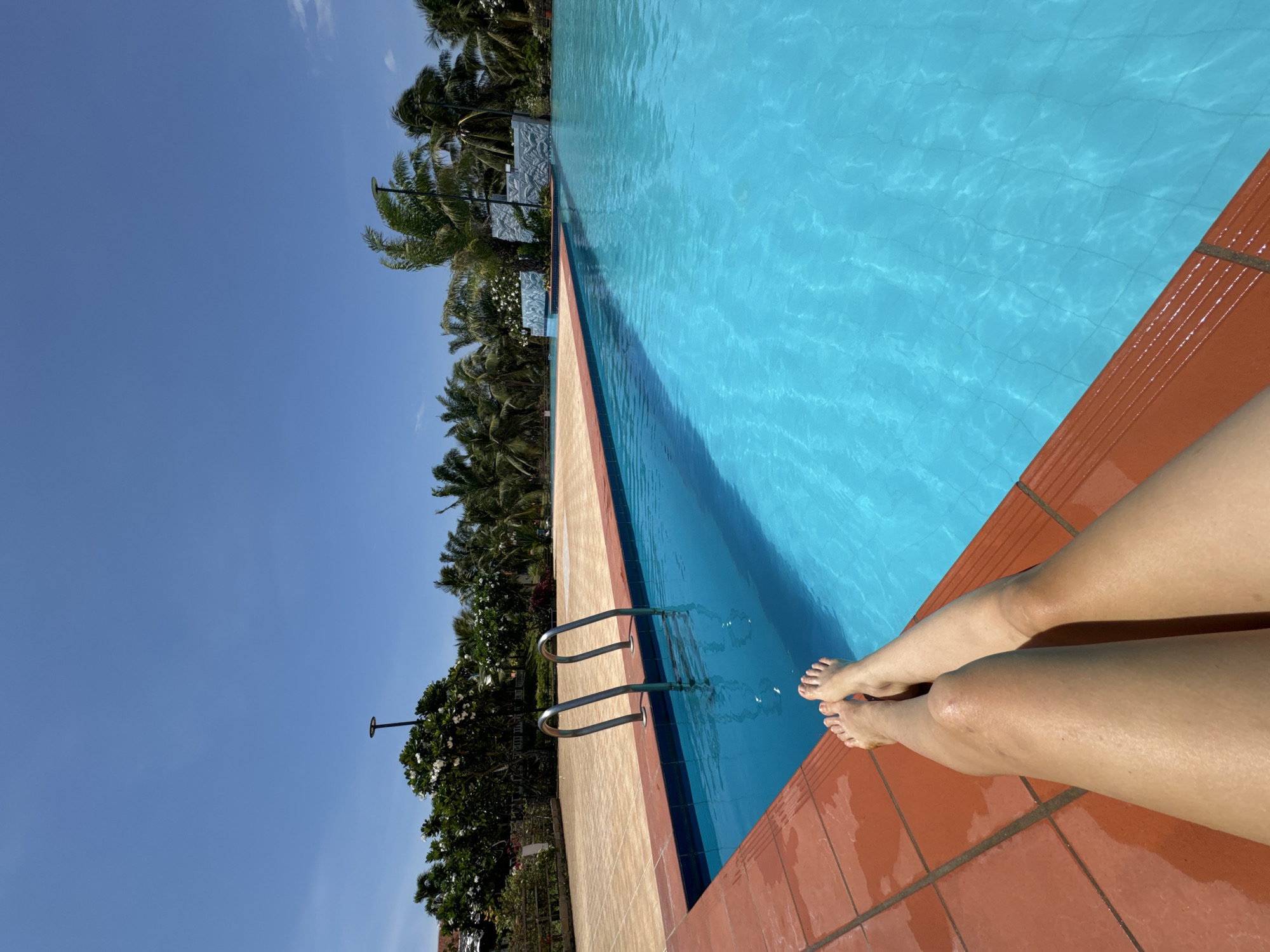 A slightly tilted outdoor shot depicts a person sitting by a swimming pool in Phu Thuy Ward, Vietnam. The pool is filled with clear blue water and surrounded by tan and reddish-brown tiles. A metal ladder is visible inside the pool. The person's legs and feet are visible resting on the tiles. Palm trees and other greenery are situated along the side of the pool, with a clear blue sky above. A lamp post is seen among the trees.