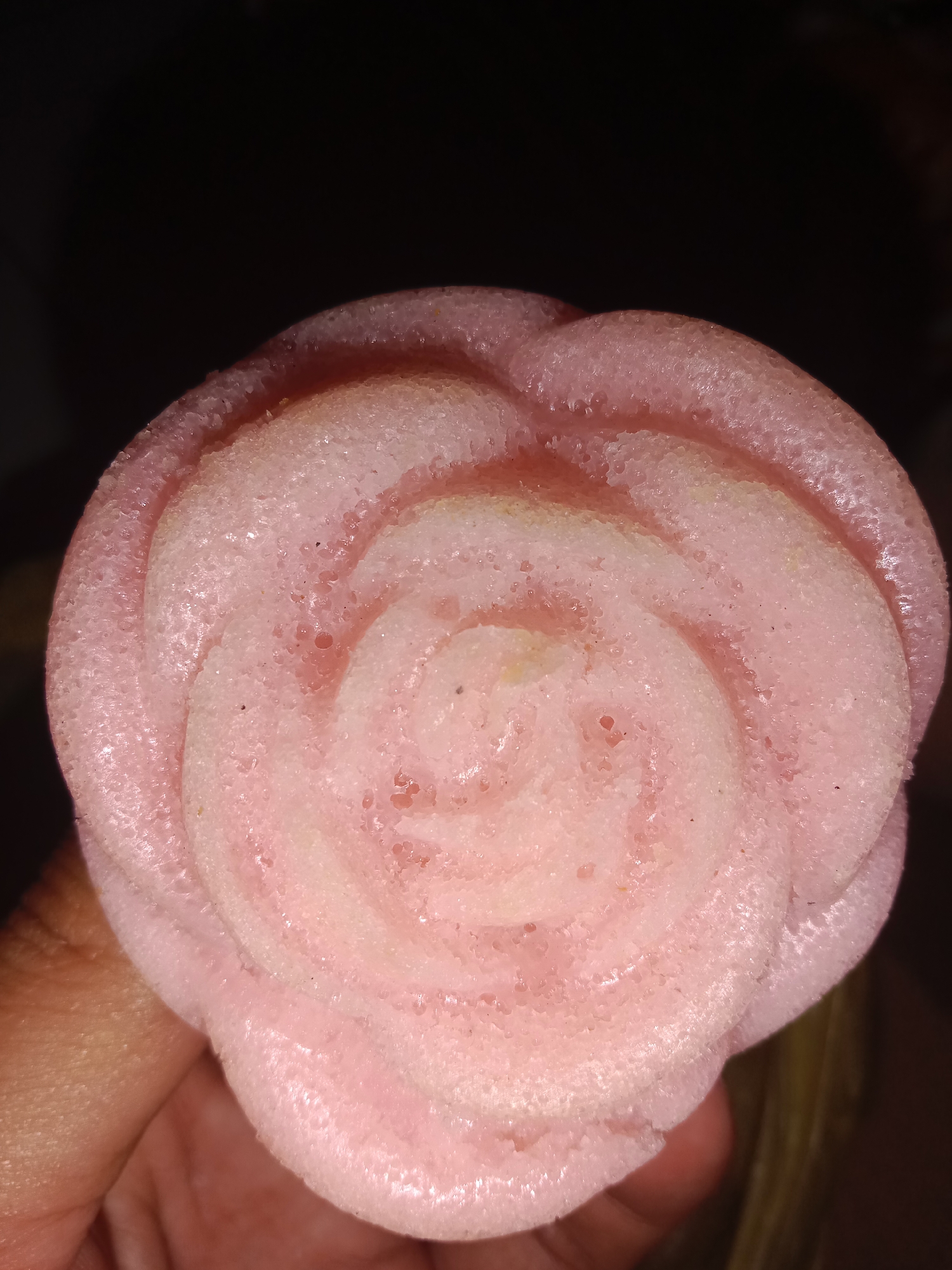A close-up, top-down shot shows a light pink, rose-shaped sweet bread held by a person's fingers against a dark background. The bread has a textured, porous surface with visible small indentations and a slight sheen, indicating a moist texture. The petals are clearly defined, creating a spiral pattern from the center outward. The lighting casts a soft glow on the bread, highlighting its delicate pink hue and intricate design. The background is predominantly black, with a slight blur suggesting it is out of focus.