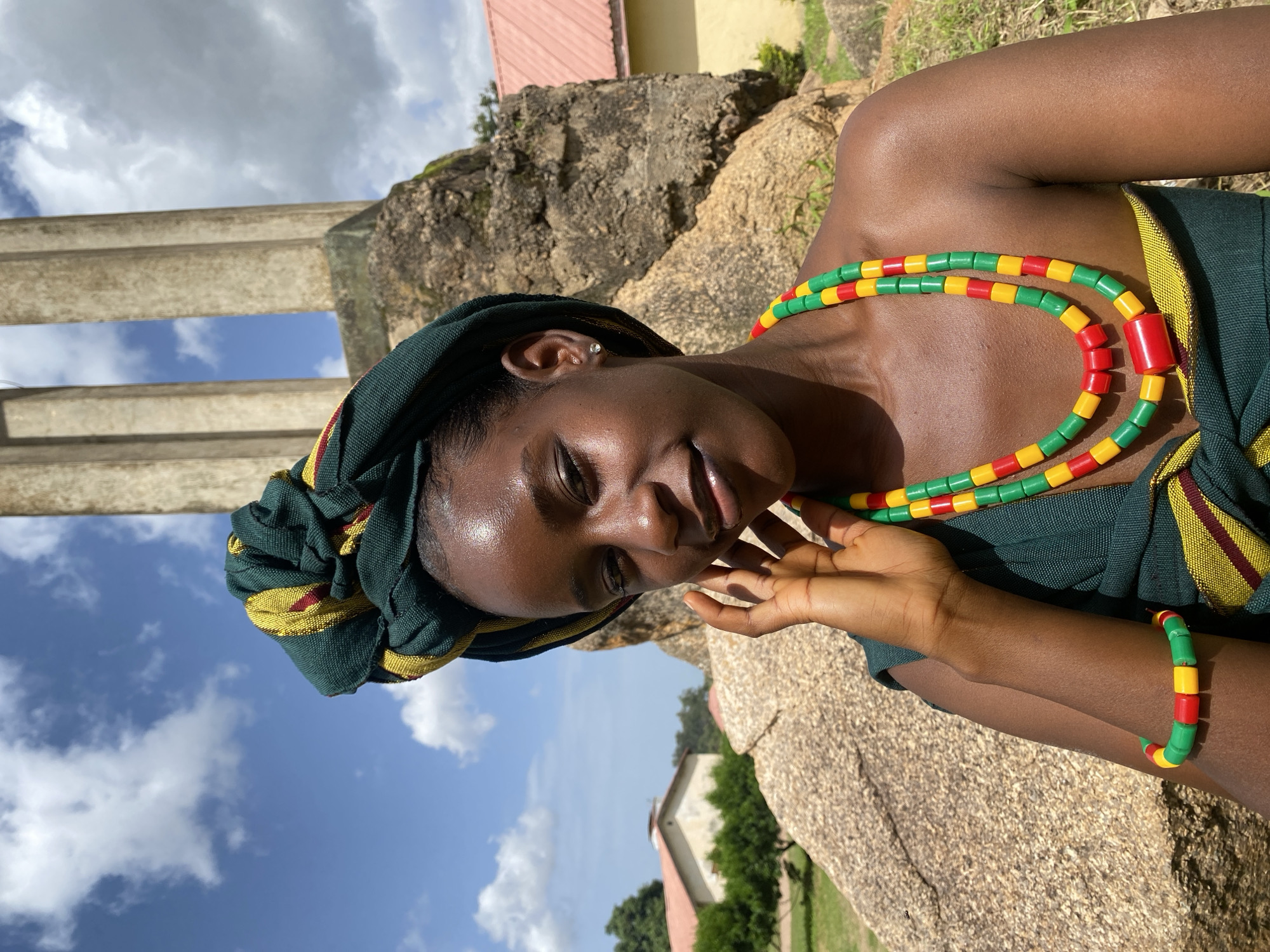 A young Black woman with dark skin and braided hair is shown in a close-up, tilted shot. She is wearing a green headwrap with yellow and red accents, and a matching green garment. She adorns herself with a beaded necklace and bracelet made of red, yellow, and green cylindrical beads. Her left hand is gently touching her cheek. She has a subtle smile, and her eyes are looking downwards, suggesting a relaxed and content emotion.
The scene is outdoors with a backdrop of large rocks and some greenery. In the distance, a building with a red roof is visible, along with trees. The sky is a vibrant blue with scattered white clouds, indicating it is daytime and likely a pleasant weather day. The lighting suggests it is likely late morning or early afternoon due to the quality of the sunlight. The location is identified as Vom, Nigeria. No text is visible in the image.