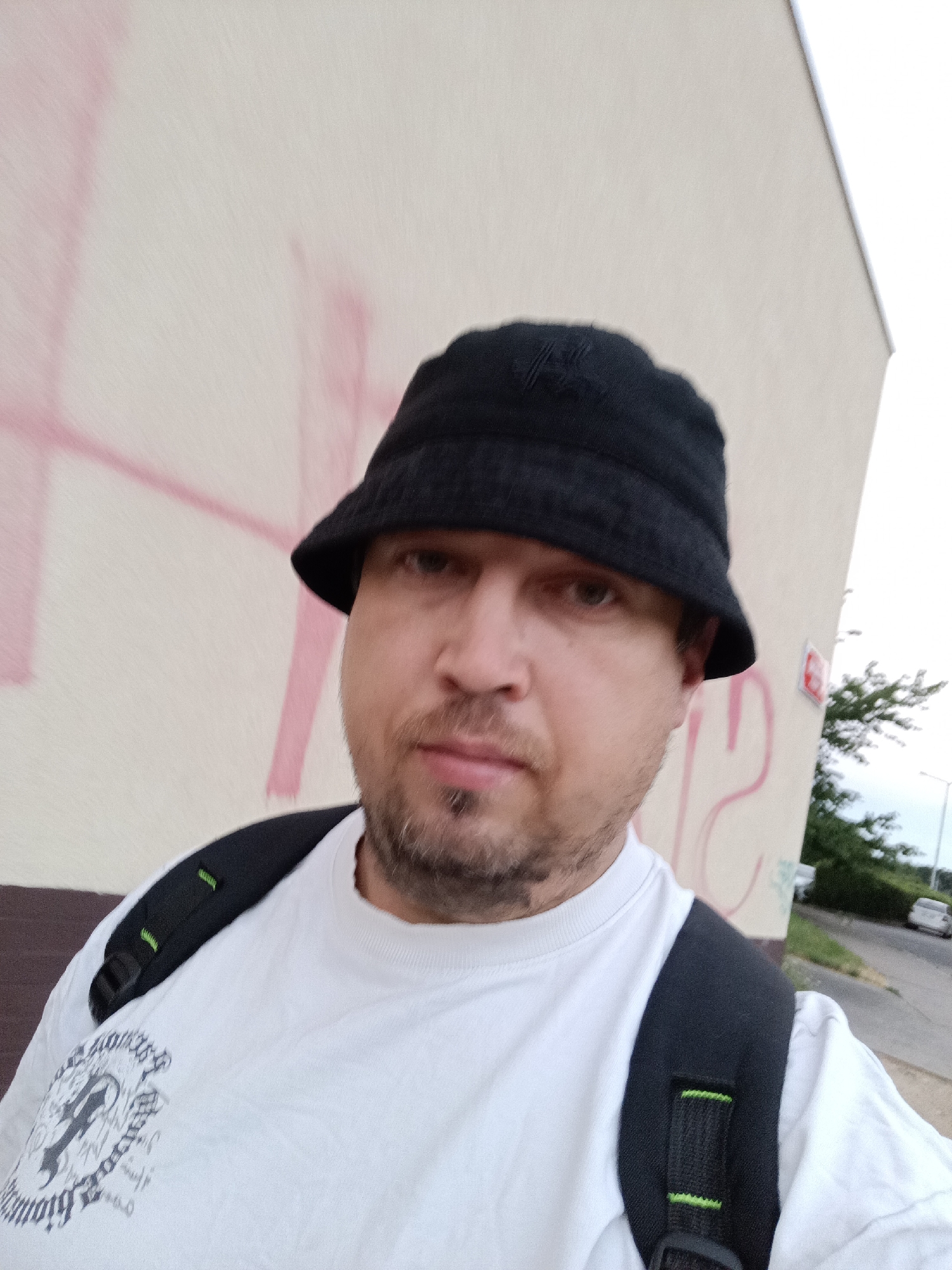 A man wearing a black bucket hat and a white t-shirt with a graphic design is standing outside against a building with pink markings. He has a backpack on, and the background shows a street with some trees and a car. The image appears to be a selfie taken in Prague, Czechia.