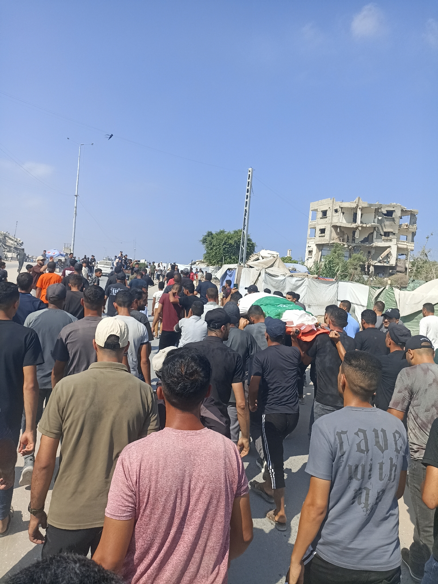 A funeral procession is underway in the city of Gaza, Palestinian Territories. A group of men are carrying a coffin draped with a green, white, and red flag, which appears to be the Palestinian flag. The procession is moving down a street lined with damaged buildings and temporary shelters. The sky is clear and blue. The people in the procession are dressed in casual clothing, with many wearing black. The atmosphere appears somber and respectful. In the background, a multi-story building is visible, showing significant damage to its structure, with several floors appearing to have collapsed or been destroyed. Tents and makeshift structures are also present, suggesting a displacement or encampment. A lamppost is also visible, and a kite is flying in the sky.