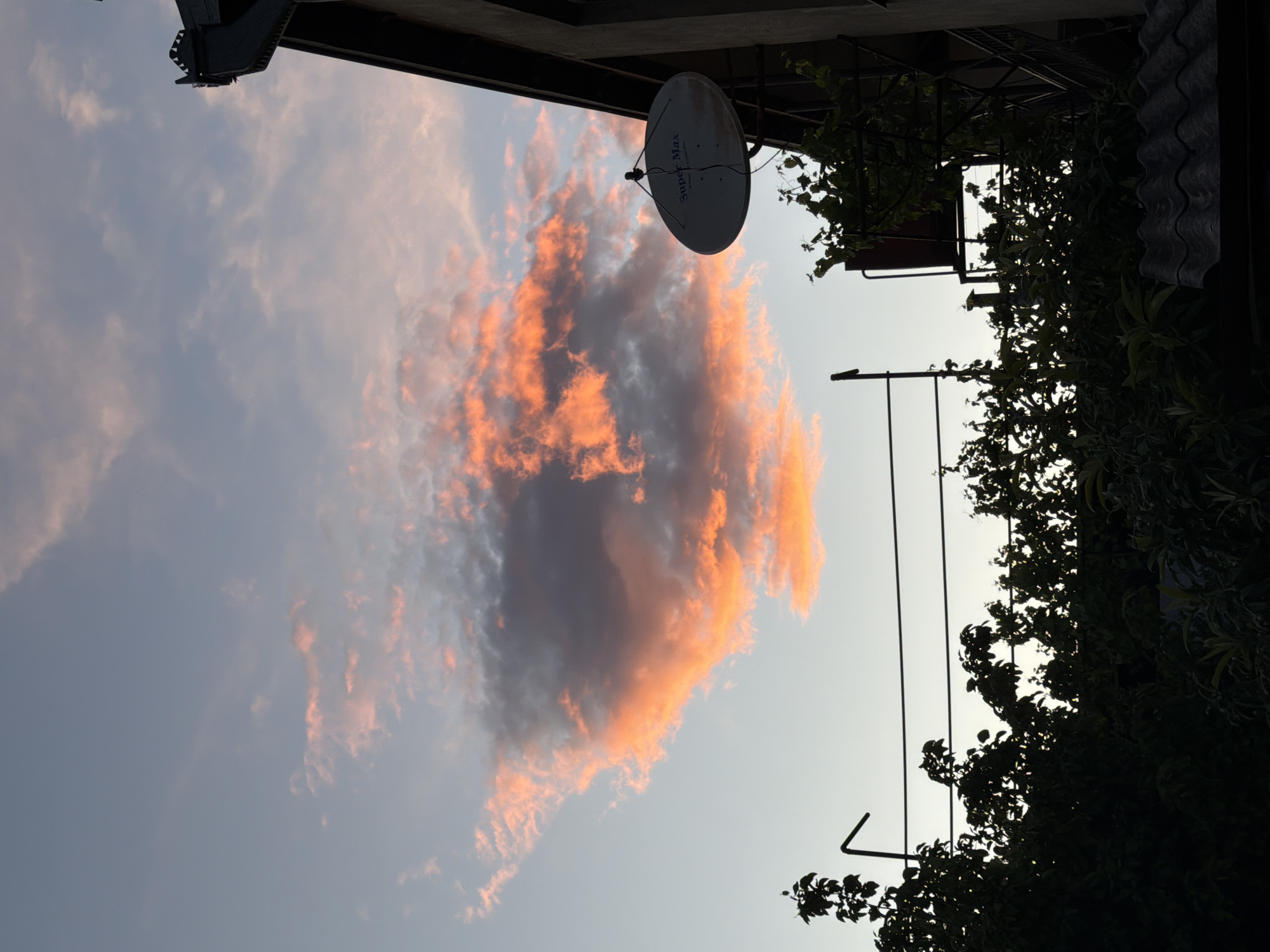 The image shows a sky in Tbilisi, Georgia. A cloud with pink edges is situated next to a white satellite dish, which is mounted on a building. The building's roof is visible at the top left of the frame. In the right part of the image are silhouettes of trees and a structure with metal bars. The picture appears to have been taken in a residential area during the evening or early morning, as indicated by the soft light.