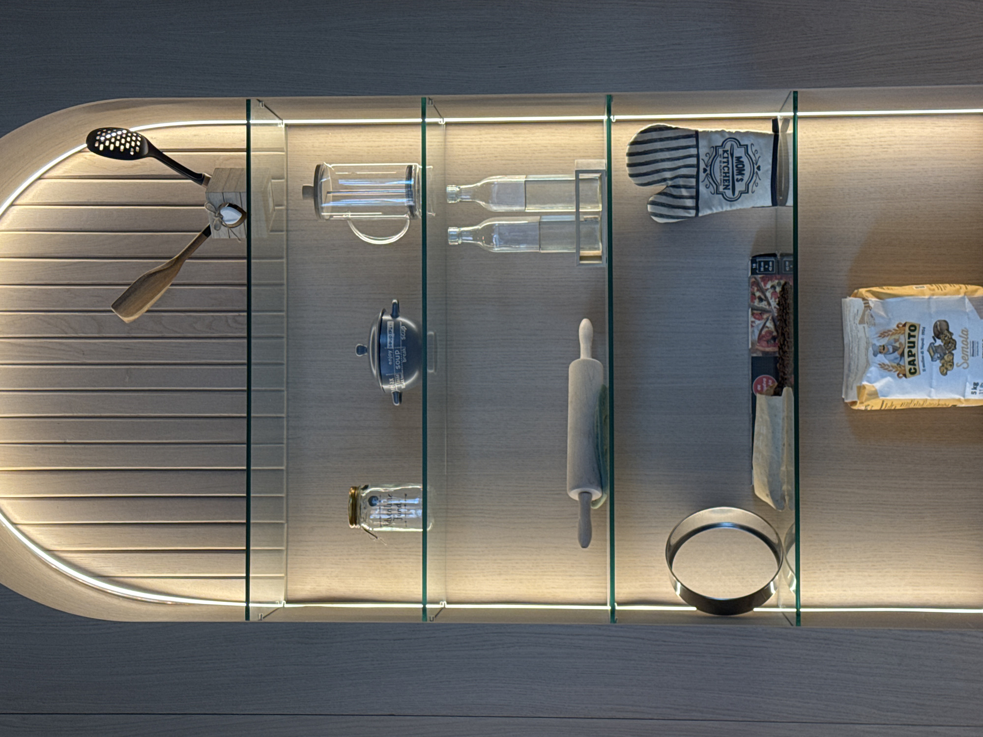 The image depicts a display cabinet containing kitchen utensils and food items. The cabinet is divided into five sections by glass panels and illuminated by LED strips. The items in the cabinet, from left to right, include: a slotted spoon, a wooden spatula, a coffee press, two glass bottles in a metal rack, a small pot, a small glass jar, an oven mitt, a rolling pin, a box of pasta with a pizza image, a cloth bag, a metal ring, and a bag of Caputo flour. The background of the cabinet is a light-colored wood. The location is likely a kitchen in Belgrade, Serbia, given the presence of Serbian language products in the image.