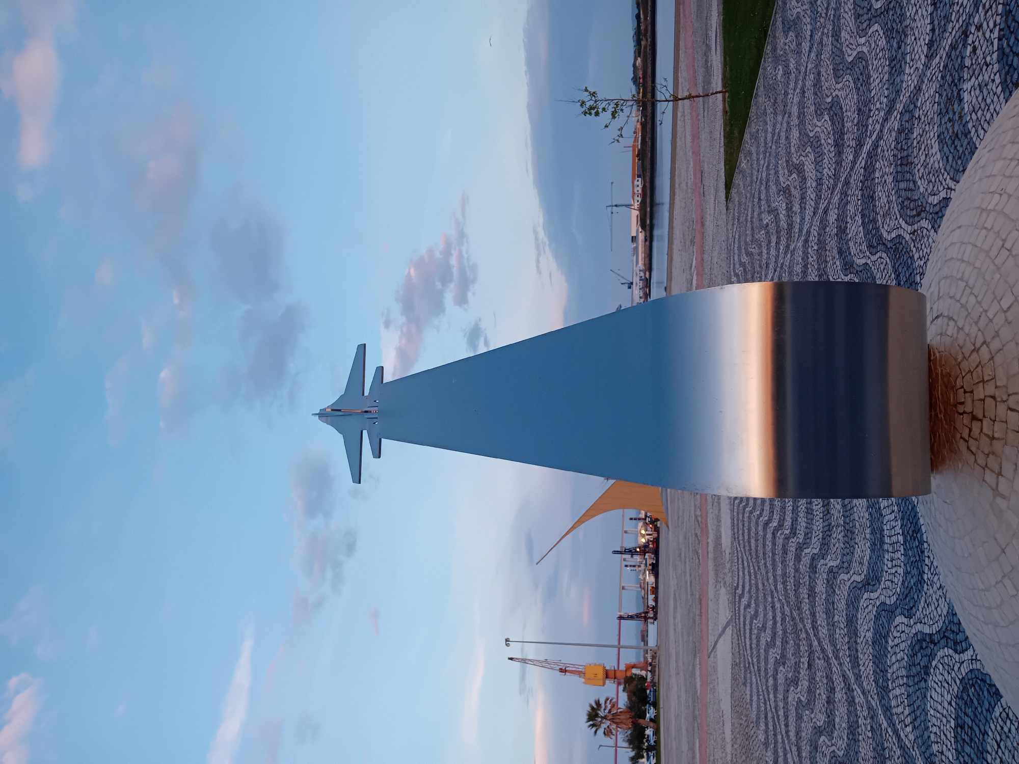 The image shows the F-5 Freedom Fighter sculpture in Figueira da Foz, Portugal. The sculpture features a stylized, metallic F-5 fighter jet atop a rectangular base, oriented towards the sky. The background reveals a mosaic-patterned walkway, a coastal area with cranes and port facilities, and a sky with scattered clouds. The lighting suggests either dusk or dawn.