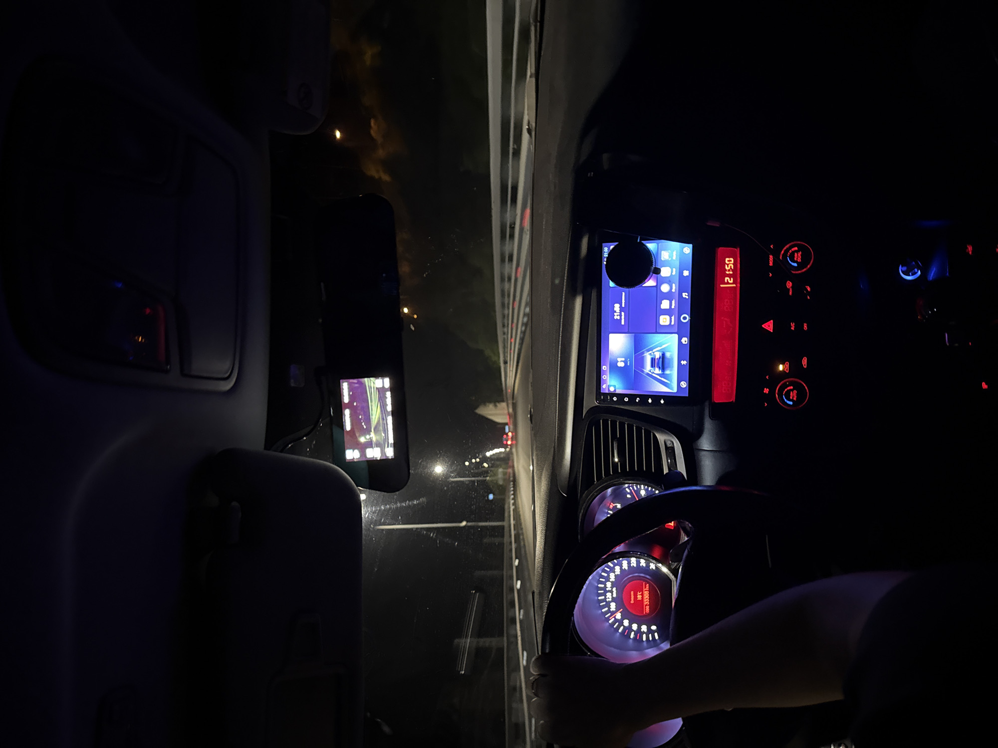 An interior view of a vehicle, likely a Kia Optima based on the dashboard design, is captured at night from the passenger's perspective, looking forward. A person's left arm and hand are visible on the steering wheel, indicating they are operating the vehicle. The speedometer shows a speed of approximately 80 km/h, the ambient temperature is 10°C, and the odometer reads 333081 km. The dashboard is illuminated with red and blue lights, including the instrument cluster, climate control buttons, and the hazard light switch. A large infotainment screen displays a car icon, various application icons, and the time 21:00. A separate red digital display below the infotainment screen shows 21:50. A dashcam is mounted near the rearview mirror, displaying a real-time feed of the road ahead, characterized by purple and green hues due to night vision or specific display settings. Through the windshield, a multi-lane road is visible, illuminated by streetlights. Guardrails line the right side of the road. The taillights of several distant vehicles are discernible. A tunnel or overpass structure appears further down the road. The scene is located in or around Chvizhepse, Russia.