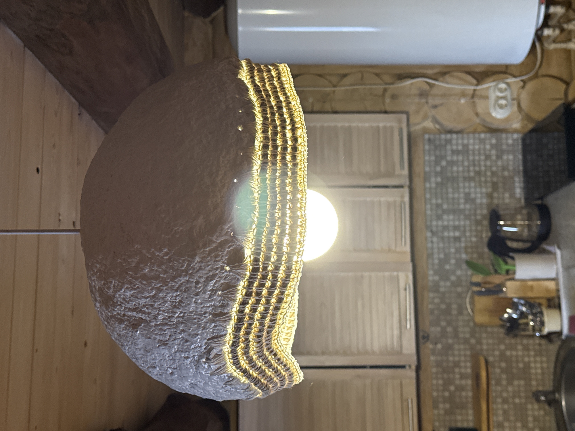 A light fixture illuminates an interior space with wooden walls. The fixture is a rounded, woven lampshade with a bright lightbulb visible through the weave. Above the lampshade, a white water heater is mounted on the wall. Below the fixture, a tiled area is visible with kitchen appliances. The location is in Tyulyuk, Russia.