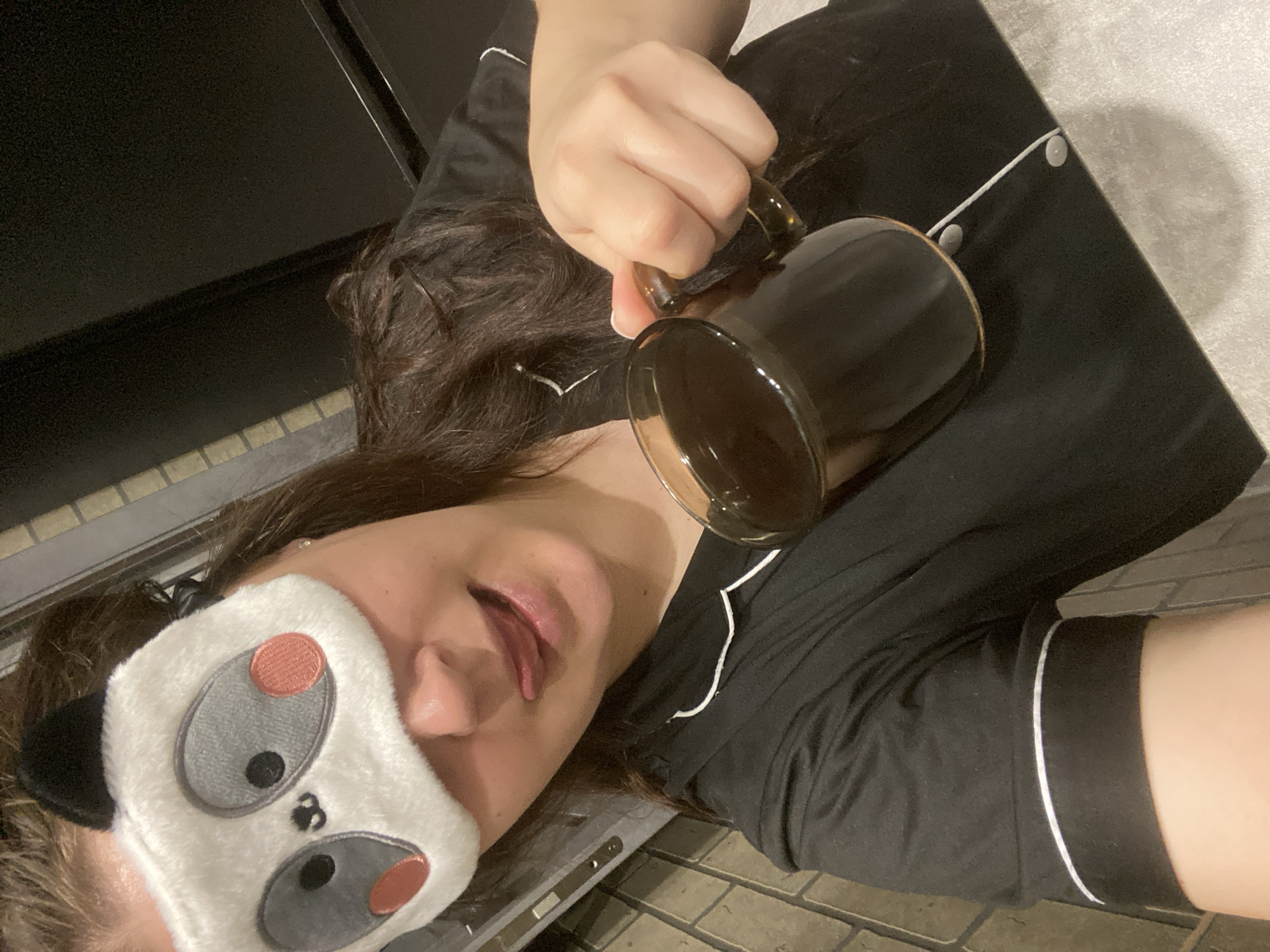 A woman wearing a panda eye mask is holding a glass mug filled with a dark liquid, possibly coffee. She is wearing black pajamas with white trim. The scene appears to be indoors, as there is what looks like a countertop or similar surface near her. The flooring visible in the image appears to be brick or tile. There is nothing to definitively indicate that this image was taken in Sochi, Russia.