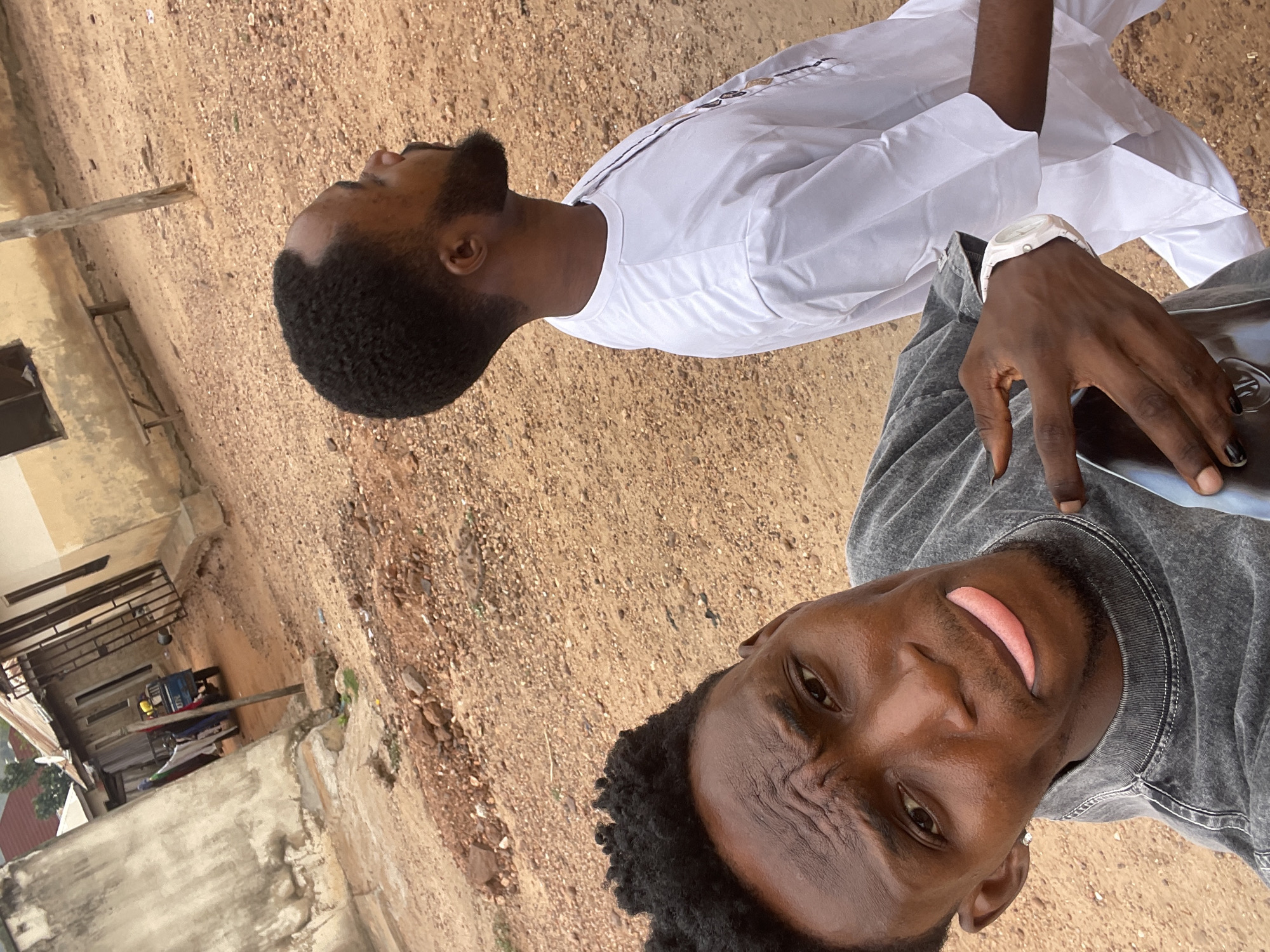 Two African men are photographed outdoors. One man wears a white, long-sleeved traditional garment. The other man is closer to the camera and wears a grey top. The man in grey is holding a dark-colored object. The ground is brown. In the background, there is a building. The location is Lokoja, Nigeria.