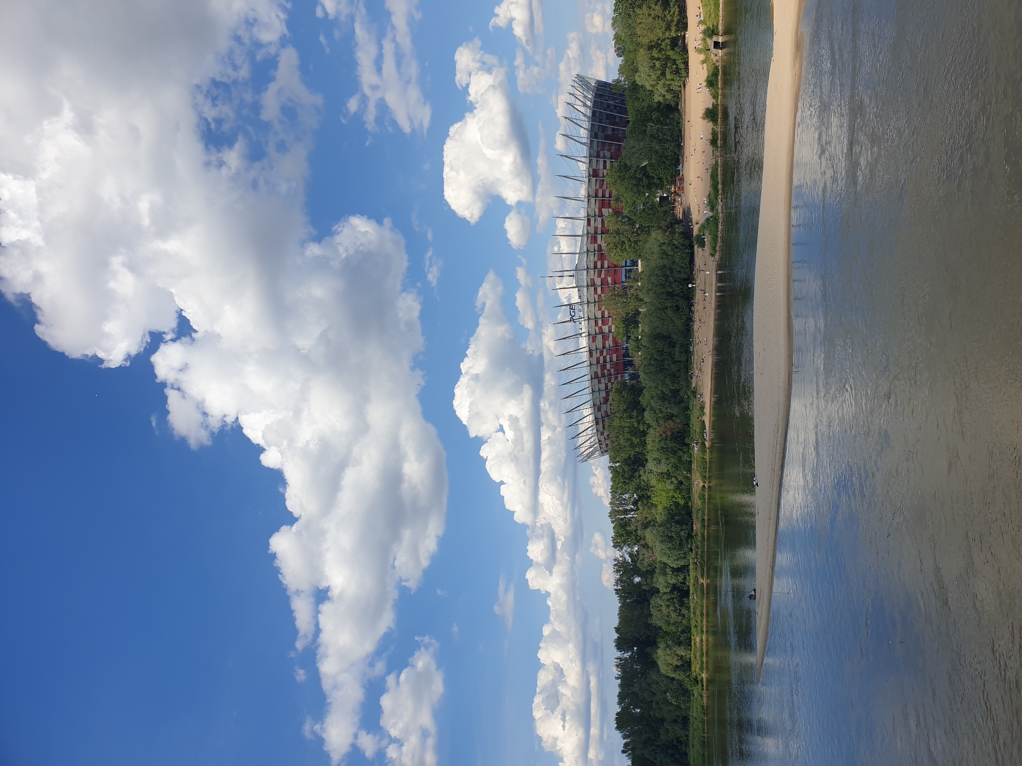 This image depicts a wide shot of the PGE Narodowy (National Stadium) in Warsaw, Poland, as viewed from the west bank of the Vistula River on a clear, sunny day. The sky occupies the upper half of the frame, featuring a vibrant blue background dotted with numerous large, white cumulus clouds.
In the middle ground, the distinctive red and white striped facade and spiky roof supports of the PGE Narodowy stadium are clearly visible, situated on the eastern bank of the Vistula. The stadium's name, "PGE NARODOWY," is legible on its upper section. A dense band of green deciduous trees lines the riverbank in front of the stadium, partially obscuring its lower half and extending across both sides of the frame.
The lower half of the image is dominated by the Vistula River. In the immediate foreground, a long, narrow sandbar or sandy island extends into the river from the left, its light brown surface contrasting with the water. Two individuals are discernible on this sandbar: one seated near a small, dark, upright object (possibly fishing equipment or a rudimentary structure) towards the left, and another figure further to the right. The river water exhibits a variable greenish-blue to brownish-grey hue, with some areas reflecting the sky.
On the far bank of the river, between the water's edge and the line of trees, a wide sandy beach is visible, populated by numerous small figures of people lounging, sunbathing, and walking, indicating recreational activity.