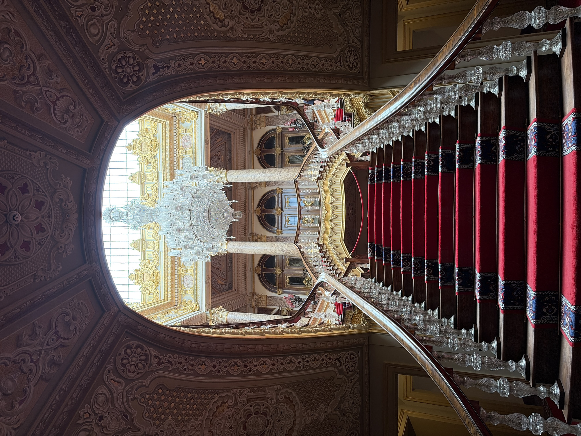 The image shows the grand staircase inside Dolmabahçe Palace, located in the Beşiktaş district of Istanbul, Turkey. The camera is positioned at the base of the staircase, looking upwards. The staircase has red carpeting with patterned borders on each step, flanked by a crystal banister. Above the staircase, a large chandelier hangs from an ornate ceiling, illuminated by natural light coming through a window. The walls and ceiling surrounding the staircase feature intricate architectural detailing. The overall environment suggests a high-end historical space.