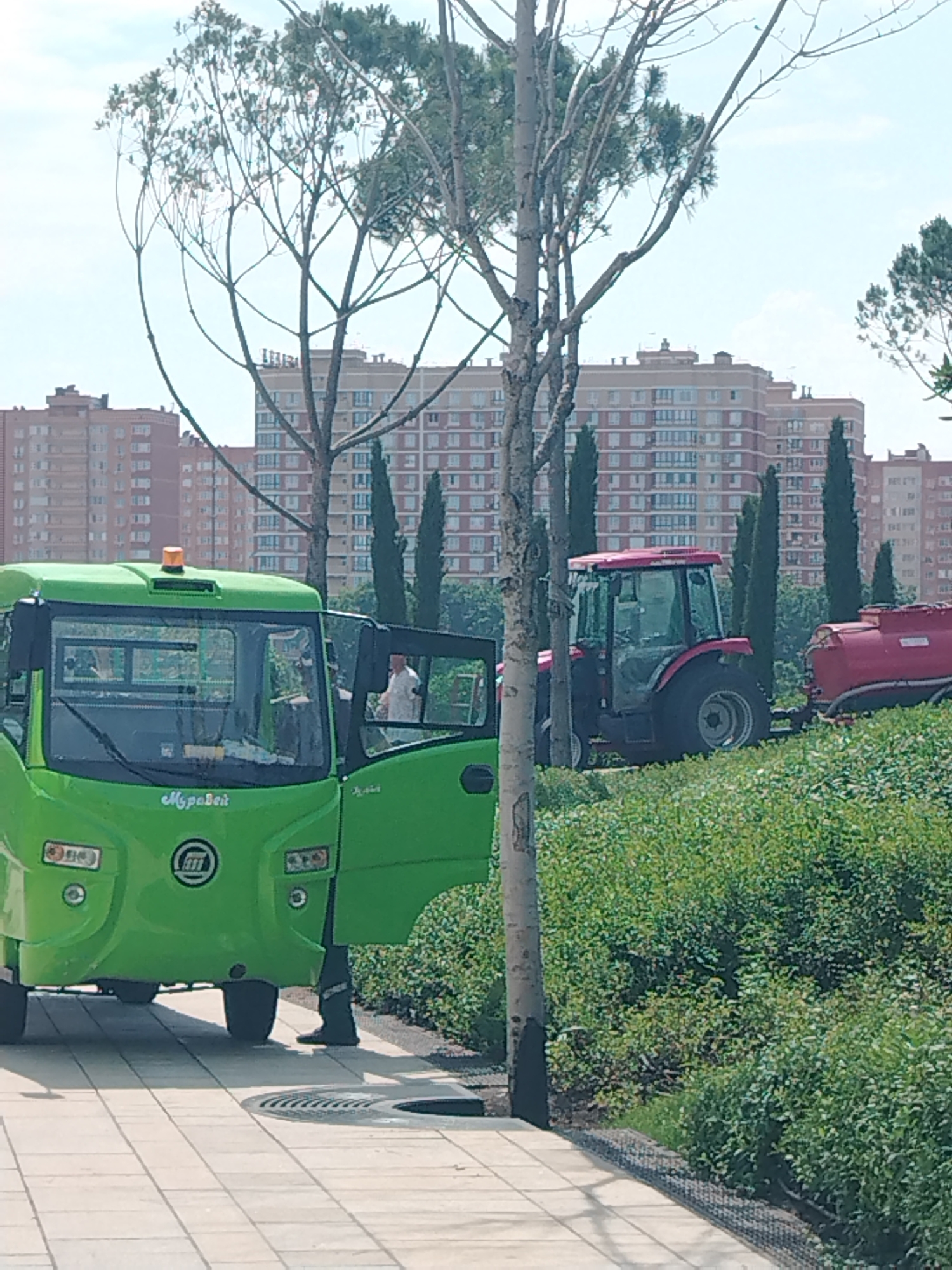 The image depicts a park or green space with urban buildings in the background, possibly in Krasnodar, Russia, given the context provided. In the foreground, a bright green vehicle, likely an electric utility cart or small bus, is parked on a paved pathway. The driver's side door of the green vehicle is open, and a person wearing dark pants and possibly a light-colored shirt is partially visible inside, appearing to be operating the vehicle or disembarking. To the right of the green vehicle, a red tractor is parked next to a red liquid storage tank. The tractor and tank are situated on a grassy embankment covered in lush green bushes. Several trees, with slender trunks and sparse foliage, are scattered throughout the scene. In the background, multi-story residential buildings with beige and red brick facades dominate the skyline. The sky is bright and appears to be overcast or hazy.