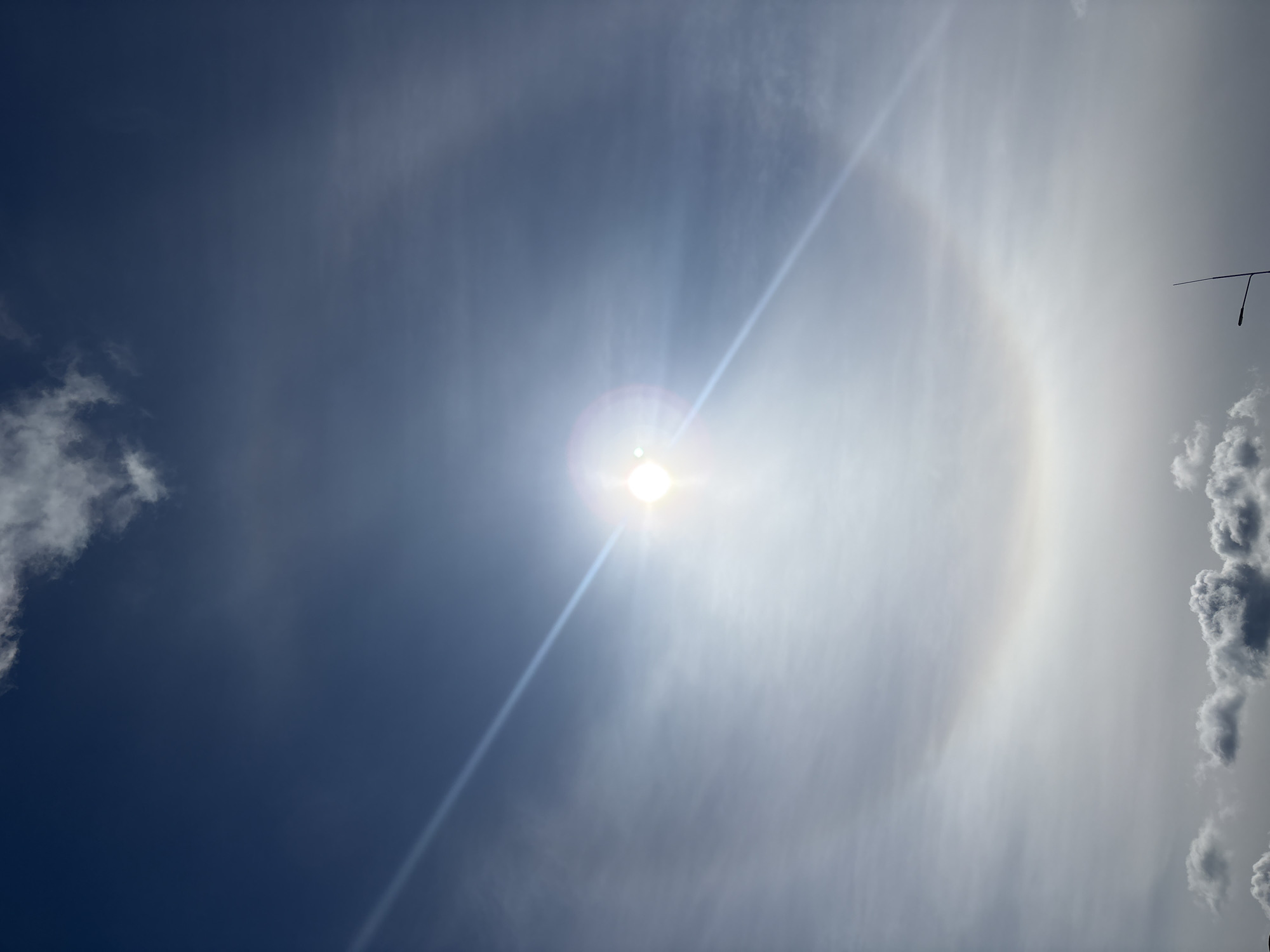 The image shows a bright sun in a blue and white sky. A 22° halo surrounds the sun. Some clouds are visible in the lower right and upper left corners of the image. A thin straight object (possibly an antenna or wire) is visible against the sky in the upper right. The location is indicated as the city of Mozhga, Russia, but no specific details in the image confirm this.