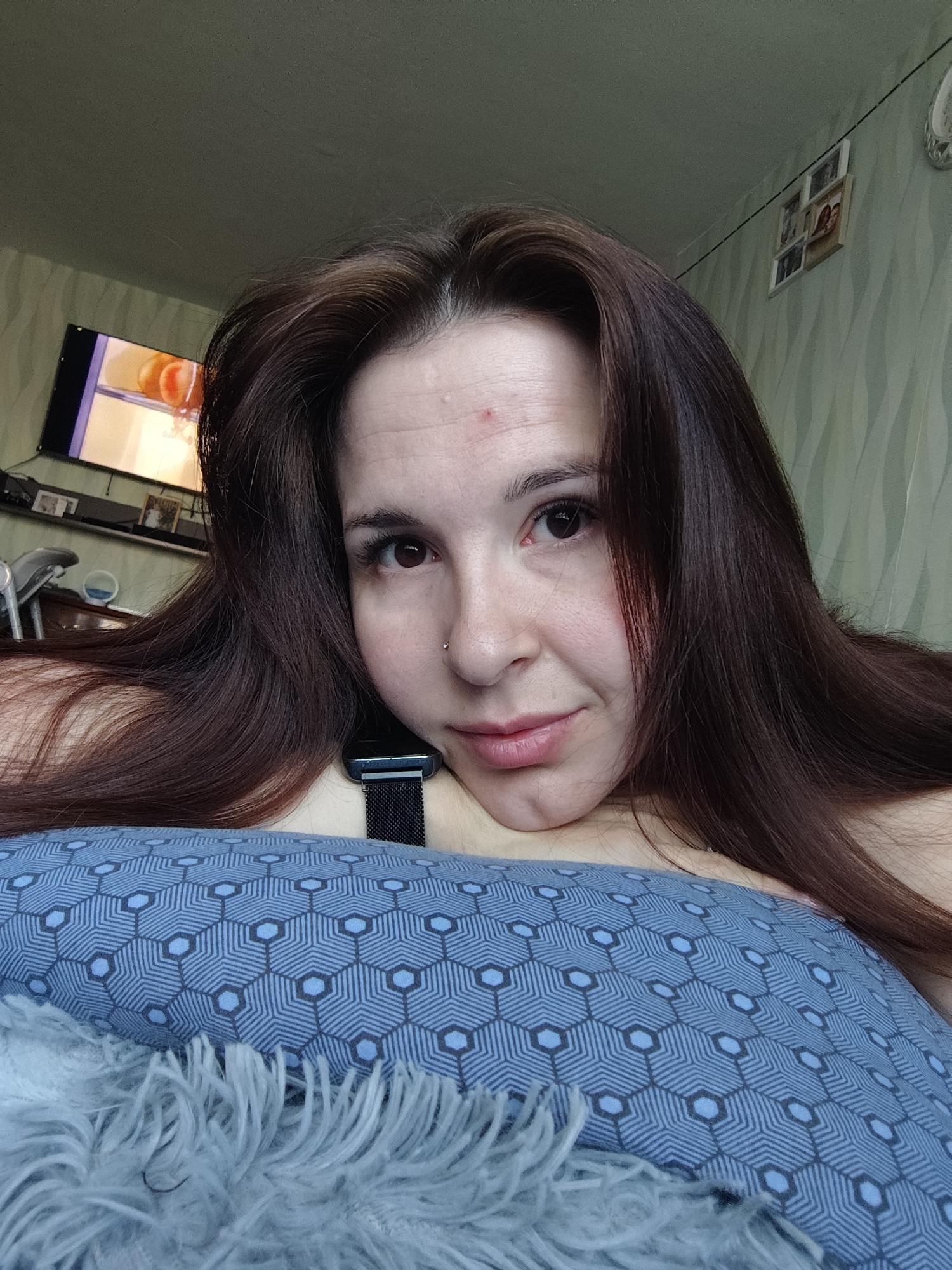 A woman is lying down, possibly on a bed or sofa, with a blue pillow beneath her. The pillow has a geometric hexagonal pattern with lighter blue accents. She has long, dark brown hair and a fair complexion. A visible blemish is present on her forehead. She is wearing a black wristwatch. In the background, a television is mounted on the wall, displaying an image with orange hues. Above the television is a shelf with various objects. A framed set of small photos is on the wall. The wall has a light green wavy pattern. The location is Фоки, Russia.