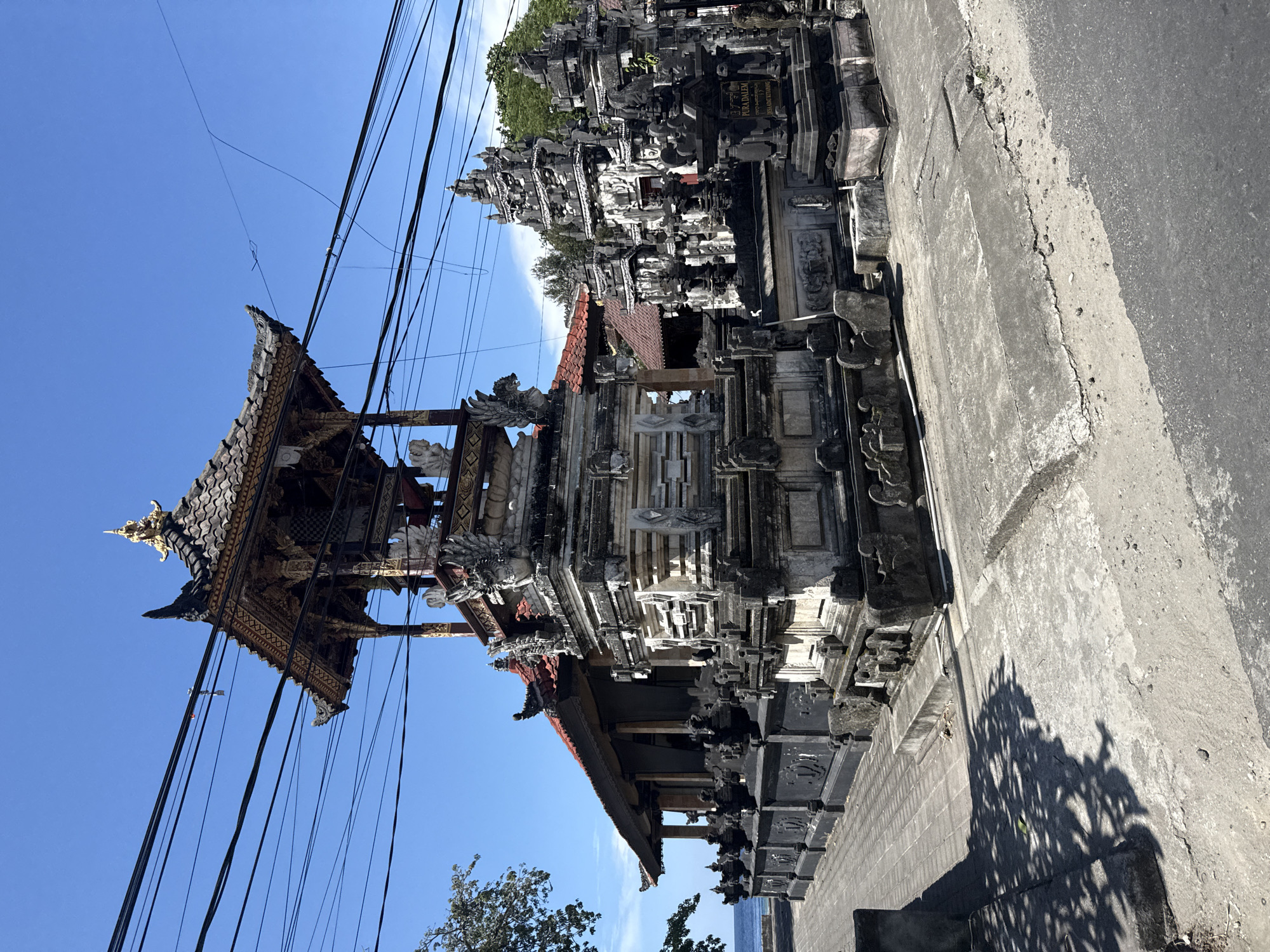 The image shows a detailed view of a traditional Balinese temple structure in Batununggal, Indonesia. The temple is characterized by its multi-tiered roof, intricate carvings, and stone construction. Power lines intersect the view of the blue sky above. The temple complex is partially obscured by adjacent buildings, with a concrete sidewalk and possible roadway visible in the foreground. Tree shadows are cast on the sidewalk, suggesting bright sunlight.