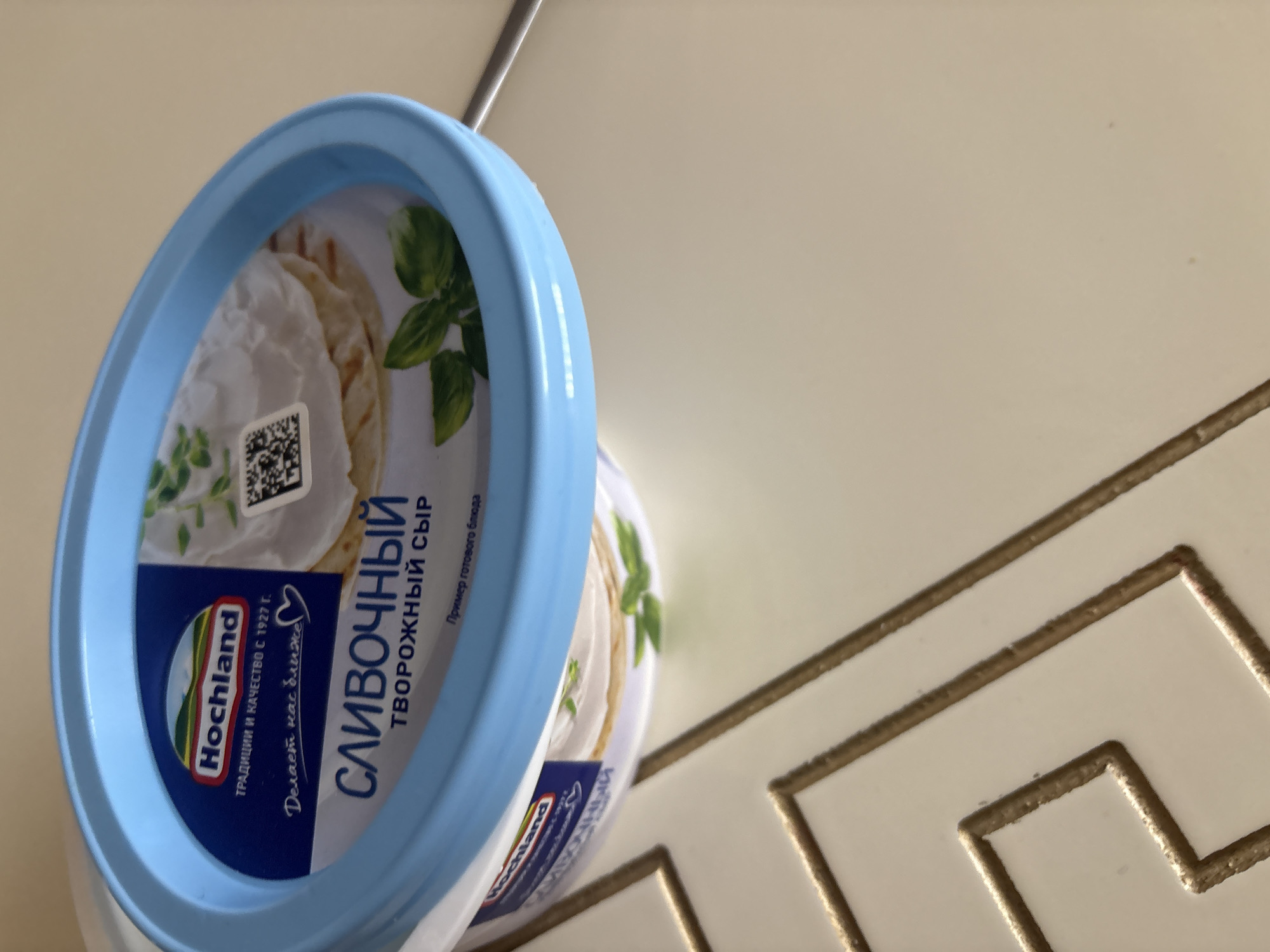 A light blue plastic tub of cream cheese is placed on a cream-colored surface with gold trim. The tub has a blue lid with the brand name "Hochland" in red and white letters. Below the brand name, there are Russian words that translate to "Traditions and Quality since 1927" and "Makes us closer," with a heart symbol. The label also shows "CREAMY" in large blue letters, followed by "CHEESE" in smaller Russian text. A QR code is visible on the right side of the label. The cream cheese itself is white and appears spreadable, with a few green leaves, likely basil, decorating the surface. The background is a plain, light-colored surface, possibly a table or countertop, with a decorative gold line pattern visible in the lower left corner. There are no people or animals present in the image. The location is not specified in the image, and therefore cannot be determined to be Georgiyevsk, Russia.