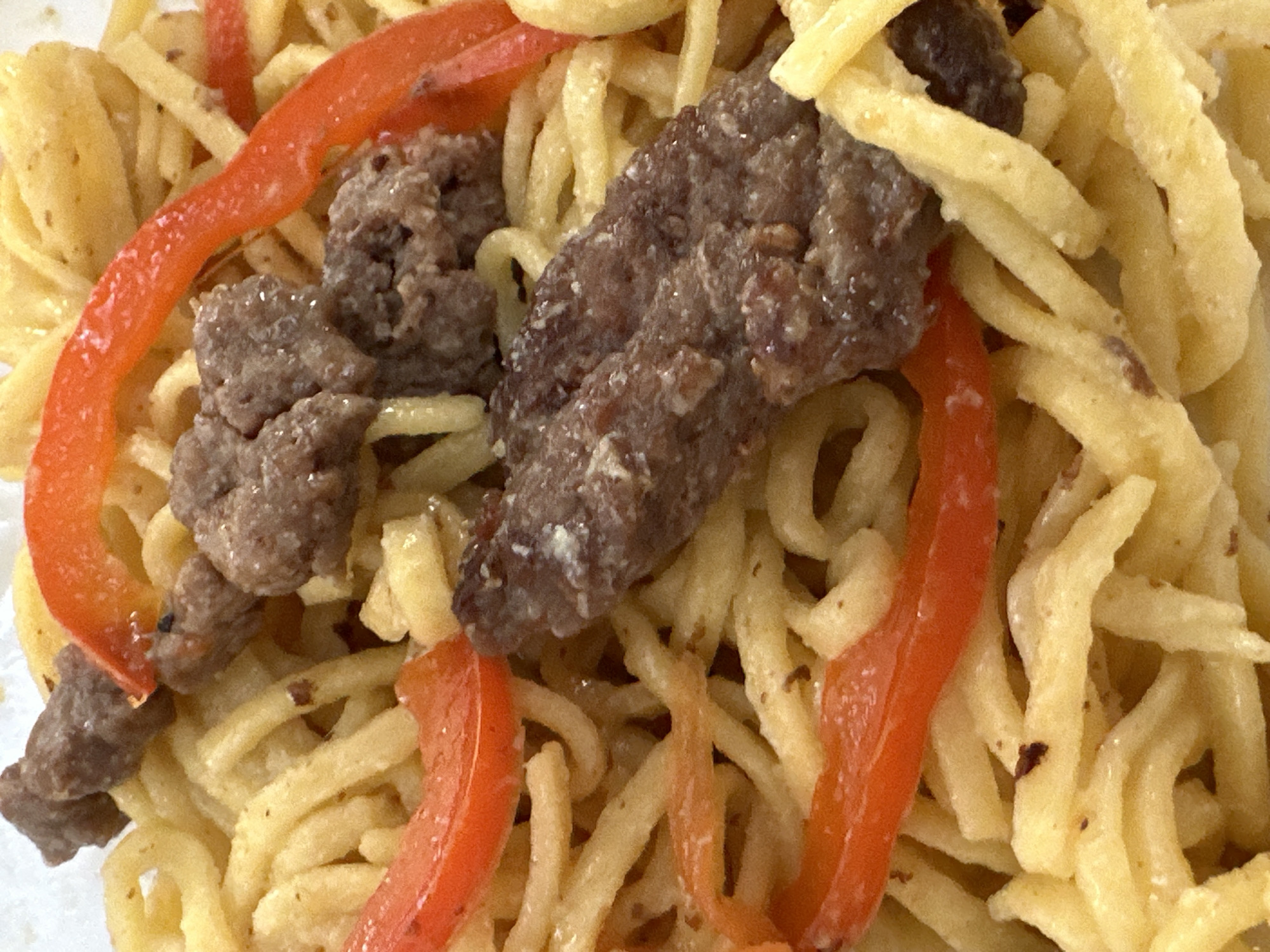 A close-up, overhead shot shows a dish of food. The food consists of a bed of light-yellow, thin, wavy noodles, interspersed with sliced red bell peppers and pieces of browned, ground meat. The meat pieces are irregularly shaped and appear to be cooked. The red pepper slices are curved and have a glossy sheen. The noodles are clustered together, with some appearing slightly oily. The overall impression is of a savory noodle dish, possibly stir-fried. There is no discernible environment outside of the food itself, and no indication of a specific city or location.