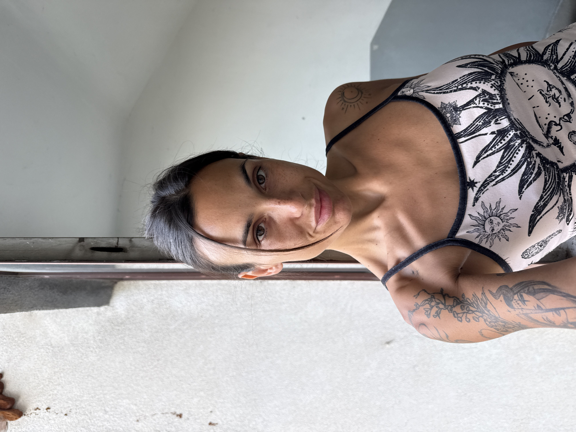 A young woman with dark hair and brown eyes poses for a selfie. She is wearing a camisole with a sun and celestial print. She has tattoos on her arms and a small tattoo on her shoulder. The background appears to be a hallway with a textured wall and a doorway. The location is identified as Pleven, Bulgaria.