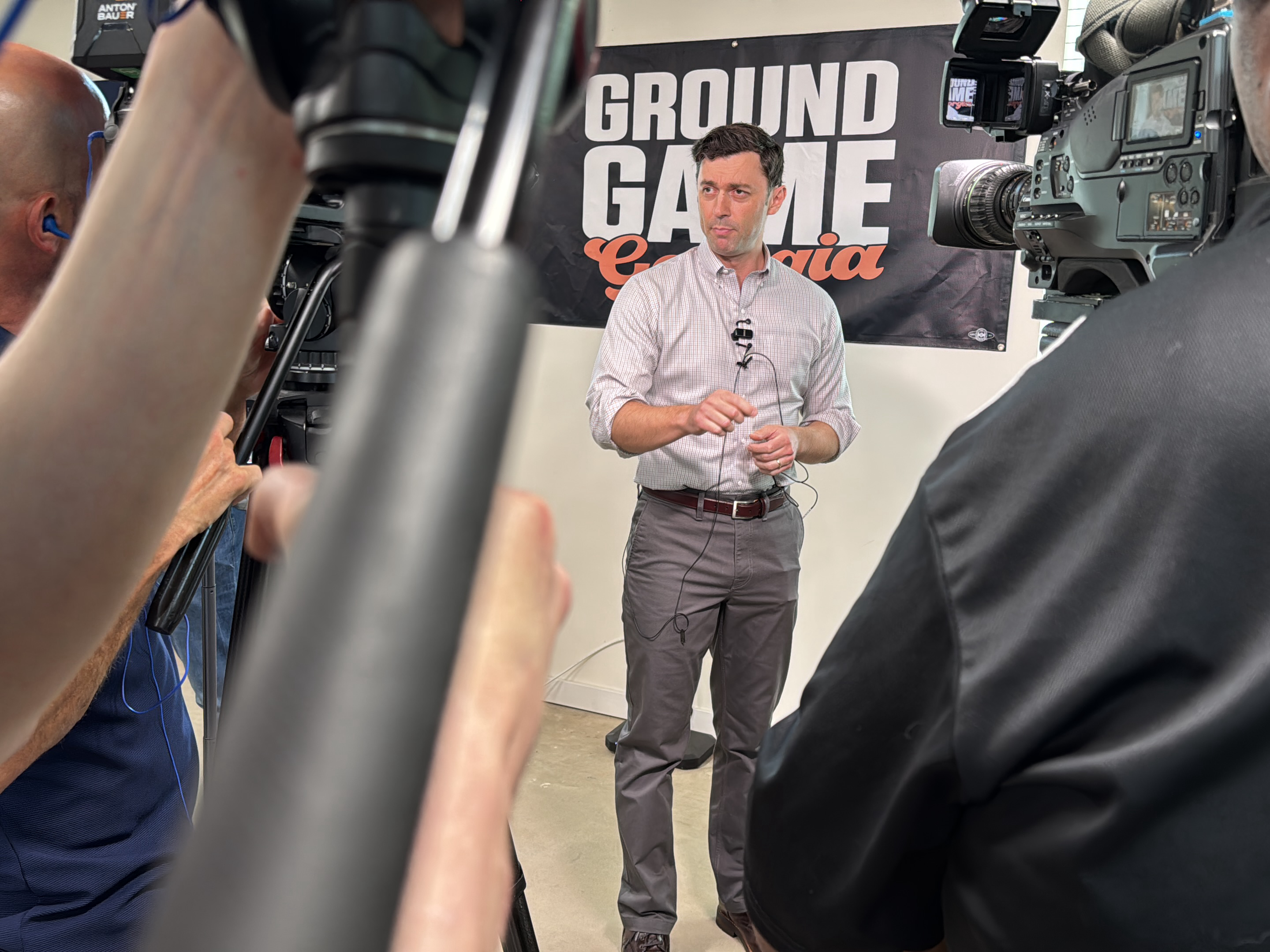 Here's a detailed description of the image:
**Scene Overview:** The image captures a man standing and speaking on camera. He is positioned in front of a banner that reads "GROUND GAME Georgia." Several cameramen surround him, obscuring parts of the view.
**People:**
* **Subject:** A man dressed in a light-colored button-down shirt and grey pants stands at the center of the frame. He wears a microphone and appears to be in the middle of a speech or interview.
* **Cameramen:** At least two cameramen are visible on either side of the subject. They operate professional-grade video cameras. One is wearing a blue shirt and the other is wearing a black jacket.
**Objects:**
* **Banner:** The "GROUND GAME Georgia" banner serves as a backdrop.
* **Cameras:** Professional video cameras are prominent, equipped with lights and viewfinders.
* **Tripods:** Camera tripods stabilize the cameras.
* **Microphone:** A lavalier microphone is attached to the subject's shirt.
* **Clothing:** The subject wears a button-down shirt, grey pants, and a belt.
**Actions:**
* The subject is speaking, gesturing with his hands.
* Cameramen are operating the cameras, likely recording the subject.
**Interactions:**
* The subject is the focus of the recording.
* Cameramen are actively engaged in capturing the footage.
**Environment:**
* The scene takes place indoors.
* The background appears to be a simple wall.
**Location:** Based on the "GROUND GAME Georgia" banner, the location may be in Georgia, United States. However, I am unable to determine the location of the city in Decatur, United States.