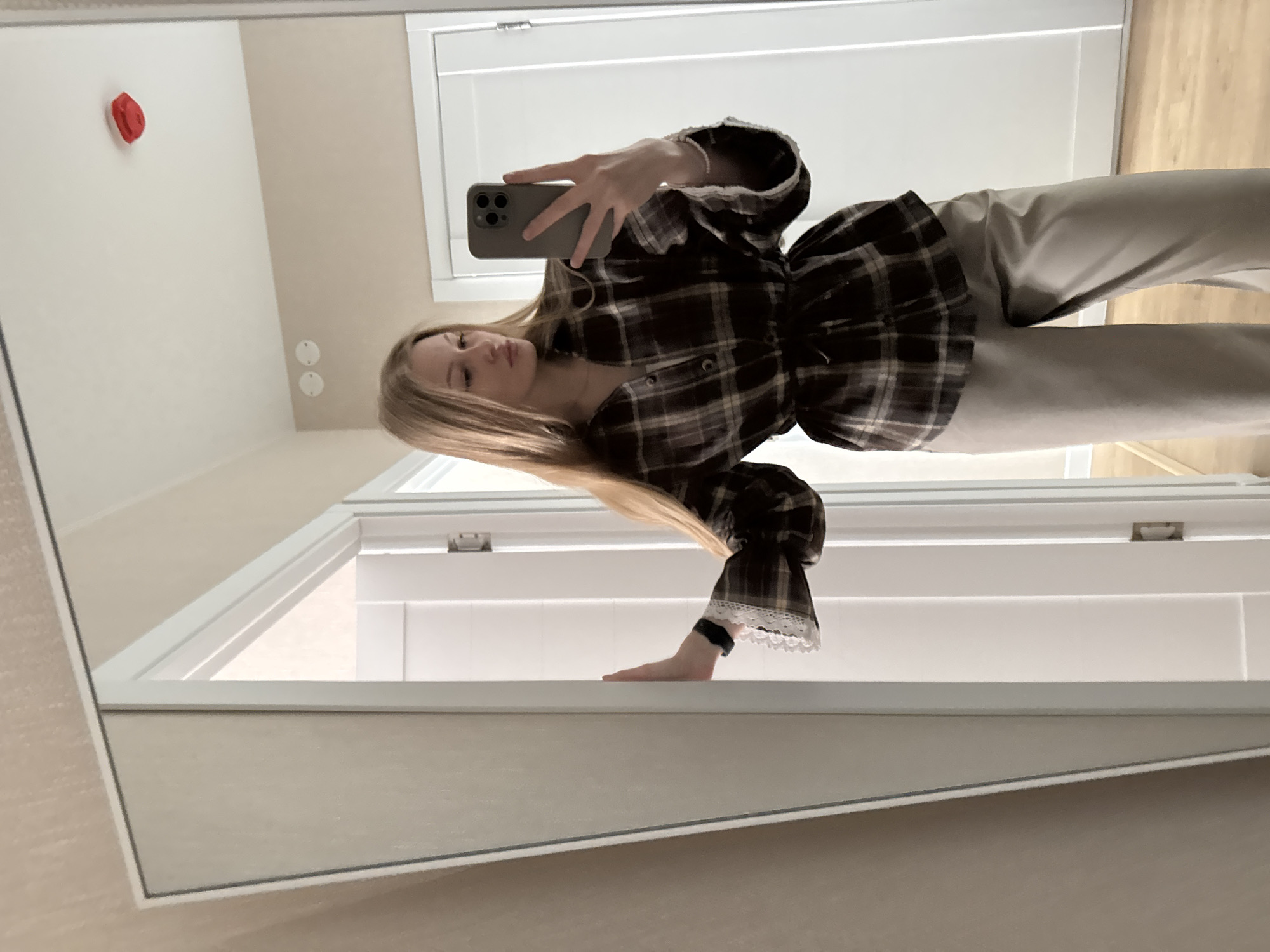 A woman in Perm, Russia, uses a smartphone to photograph herself in a mirror. She is wearing a brown plaid shirt with white lace accents on the cuffs and light-colored pants. Behind her is a white door and tan walls with a red fire alarm and light fixtures.