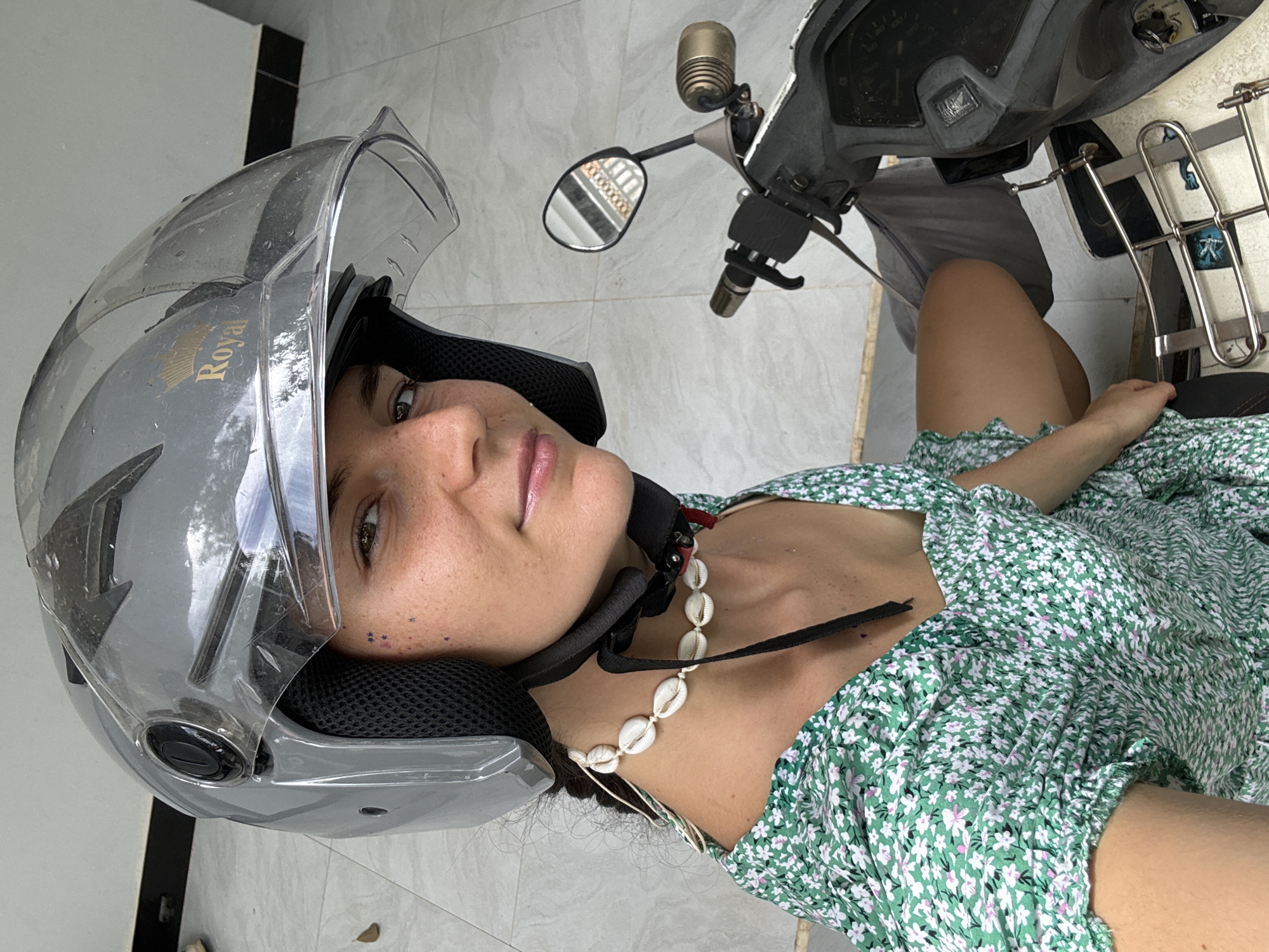 A woman wearing a silver motorcycle helmet and a green and white floral dress lies on a gray tiled surface, seemingly next to a moped. The helmet, which has the word "Royal" on it, has its visor up. The woman has fair skin and is wearing a necklace made of shells. Her moped is positioned to the right of her, with the handlebars and mirrors visible. The background suggests an outdoor or semi-outdoor area, possibly in Mũi Né, Vietnam.