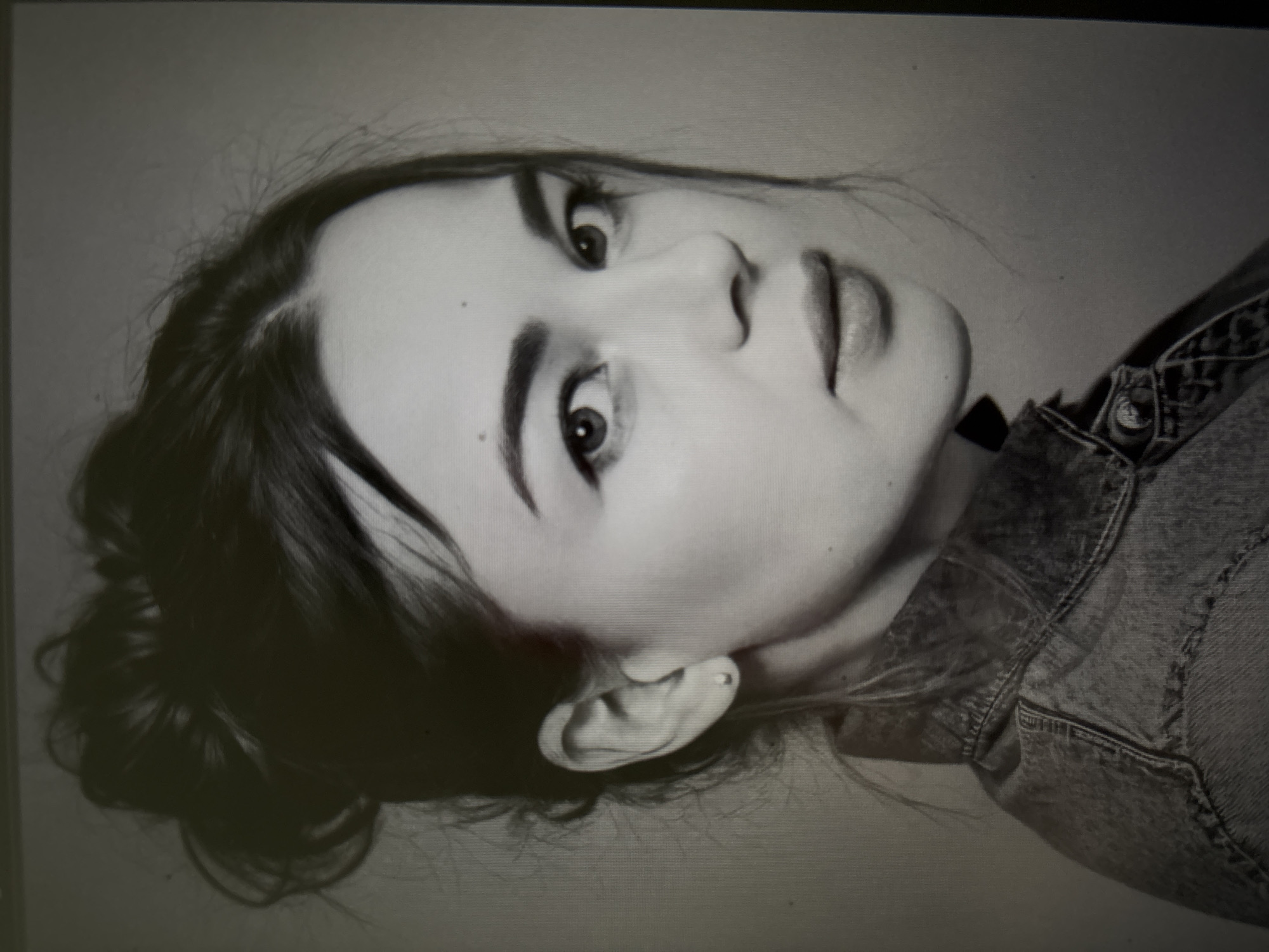 The image is a grayscale portrait of a young woman. She has her dark hair pulled up in a messy bun. She is wearing a denim jacket. Her head is turned to her right, and she is looking directly at the camera with a neutral expression. There is a faint earring visible in her left ear. The background is plain and out of focus. The image does not contain any information that would suggest a specific location in the city of Novosibirsk, Russia.