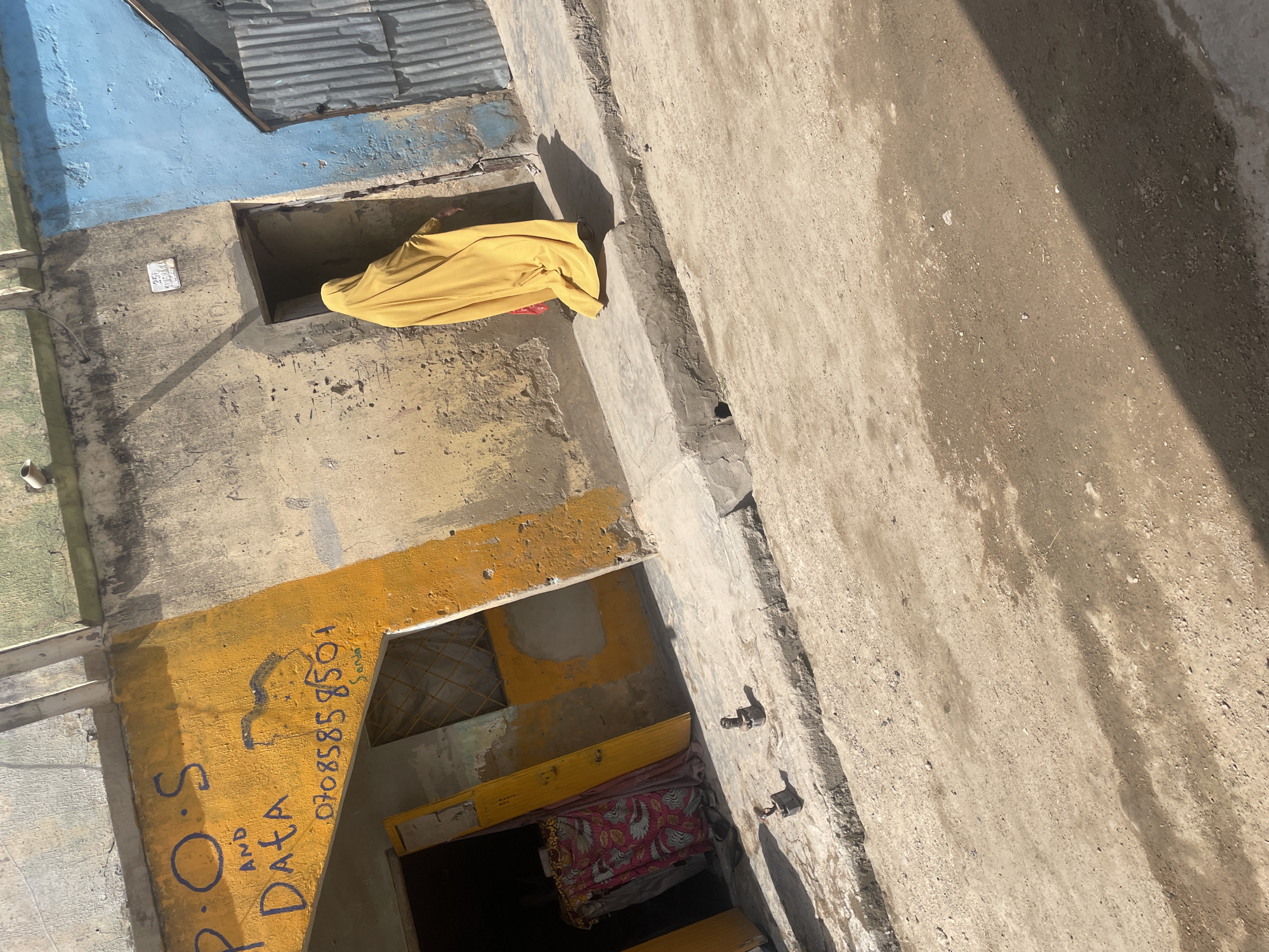 This image depicts a building in an unknown city. Part of the building's exterior is painted blue, and other parts are painted yellow. Graffiti reading "P.O.S AND DATA 07085858501" is painted on the building. A yellow cloth is draped over an opening in the building. A concrete surface is visible in the foreground.