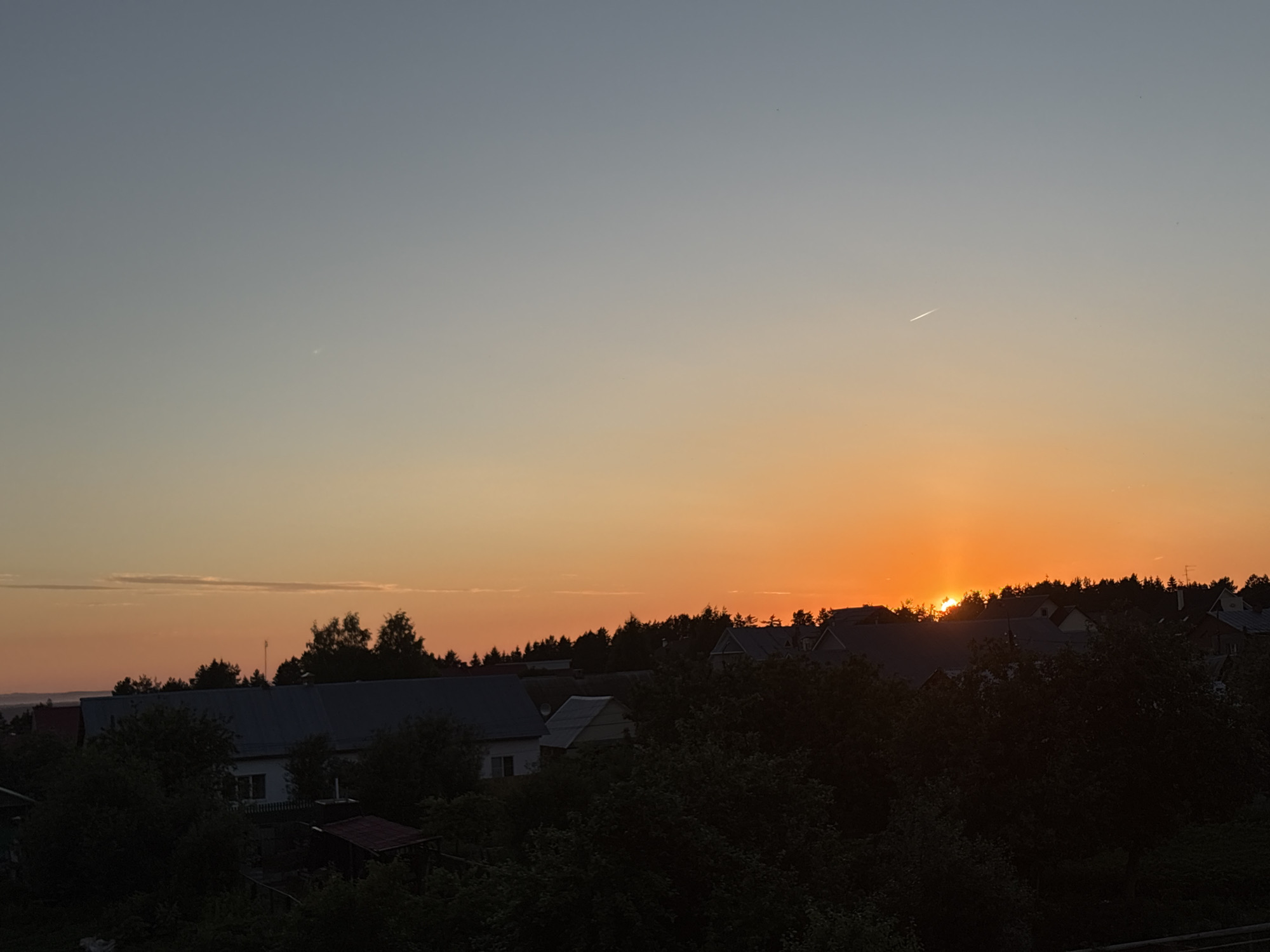 The image depicts a rural village scene in the city of Perm, Russia, during sunset. The sky transitions from a pale blue at the top to a vibrant orange and yellow near the horizon, with a faint contrail from an airplane visible in the upper sky. Silhouetted against the bright sky are the roofs of several houses and a line of trees. Dense dark foliage fills the foreground, obscuring much of the lower portion of the structures. The overall impression is of a peaceful, twilight setting in a residential area surrounded by nature.