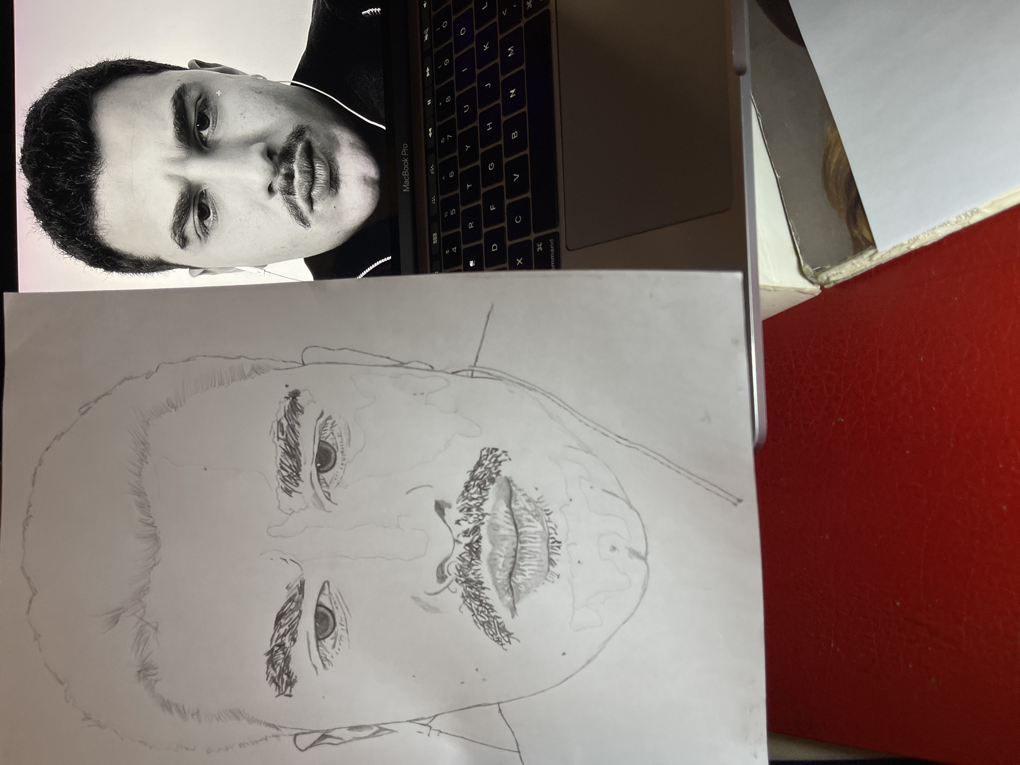 This image depicts an indoor scene in Jos, Nigeria, showcasing a pencil drawing positioned alongside a laptop displaying its reference image.
On the left, a white sheet of paper presents a monochrome pencil sketch of a male's face, appearing to be a work in progress. The drawing outlines facial features including a hairline, thick eyebrows, eyes, nose, lips, and a prominent mustache, with initial shading present on the eyebrows and mustache.
To the right, an open silver MacBook Pro laptop displays a black and white close-up portrait of a male individual on its screen. This individual has dark, short hair, thick eyebrows, and a distinct mustache. He is wearing a dark garment, possibly with a zipper closure, and white wired earphones are visible in both ears. A small white crosshair-like mark is present near his right eye (viewer's left) on the screen. The laptop's backlit keyboard and trackpad are visible below the display.
The entire setup rests on a textured, reddish-orange surface, likely a tabletop. A white-covered book, partially visible at the lower right, appears to be situated beneath the laptop, possibly providing support. The visual correspondence between the drawing and the digital image on the laptop indicates the former is being rendered from the latter.