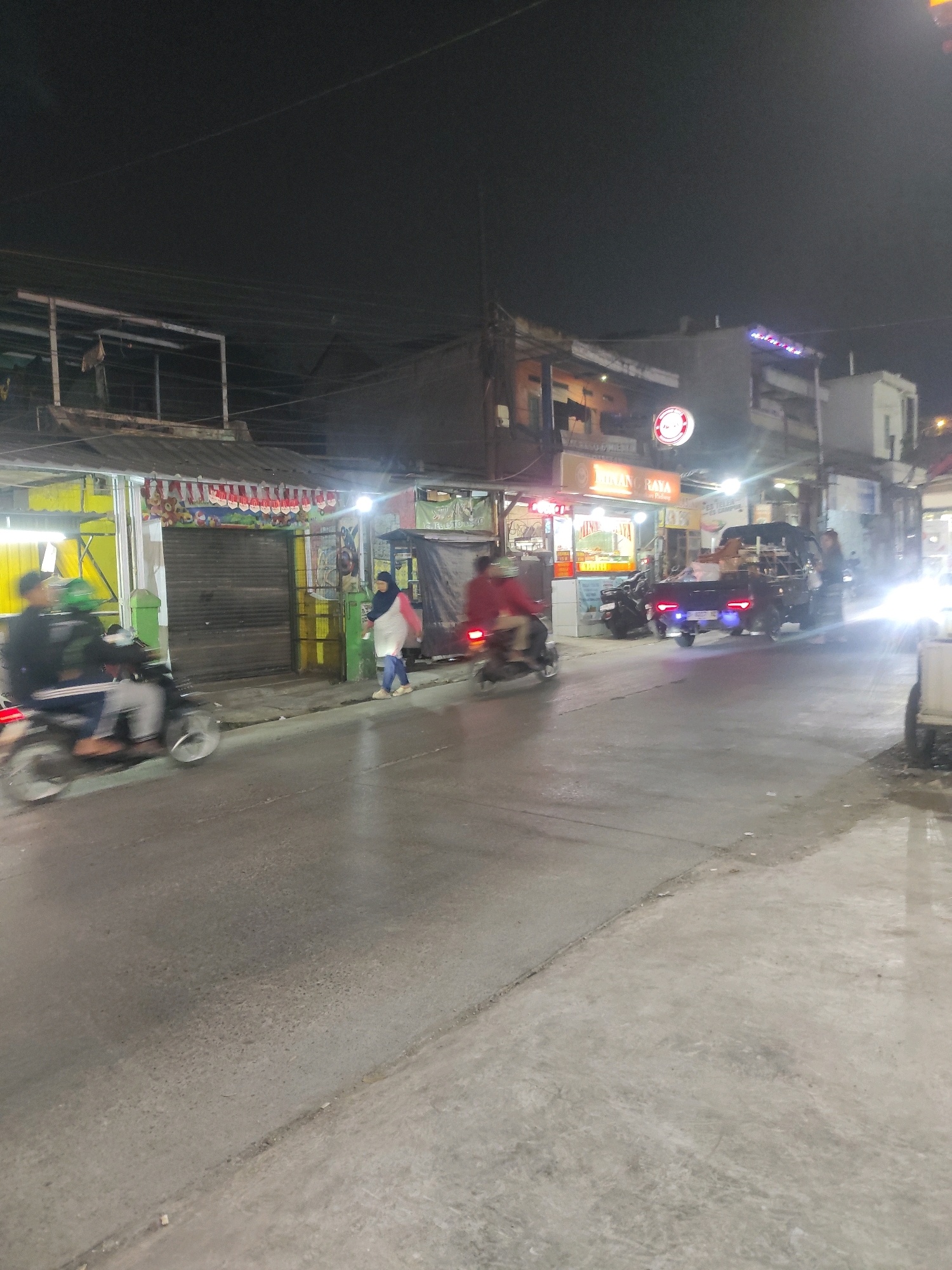 This is a nighttime, eye-level, medium shot of a street scene in Padalarang, Indonesia. The road in the foreground is paved and appears wet, reflecting the surrounding lights. On the left side of the street, there are several small businesses with open fronts, illuminated by artificial lighting. One business has a bright yellow facade and displays a row of red and white pennants overhead. A person on a motorcycle, with a green helmet and black jacket, is moving away from the camera on the left side of the road. Further down the street, to the right of the yellow-fronted shop, is a dark-colored utility vehicle. It has its headlights and taillights illuminated, and it appears to be stationary or moving slowly. A person in a white and pink top and headscarf is walking on the sidewalk in front of the shops, moving towards the right of the frame. The background consists of the facades of more buildings, some with illuminated signs. One sign displays the words "MINANG RAYA" in glowing letters, along with a circular logo. Overhead, power lines are visible against the dark night sky. The overall atmosphere is that of a busy commercial street at night.