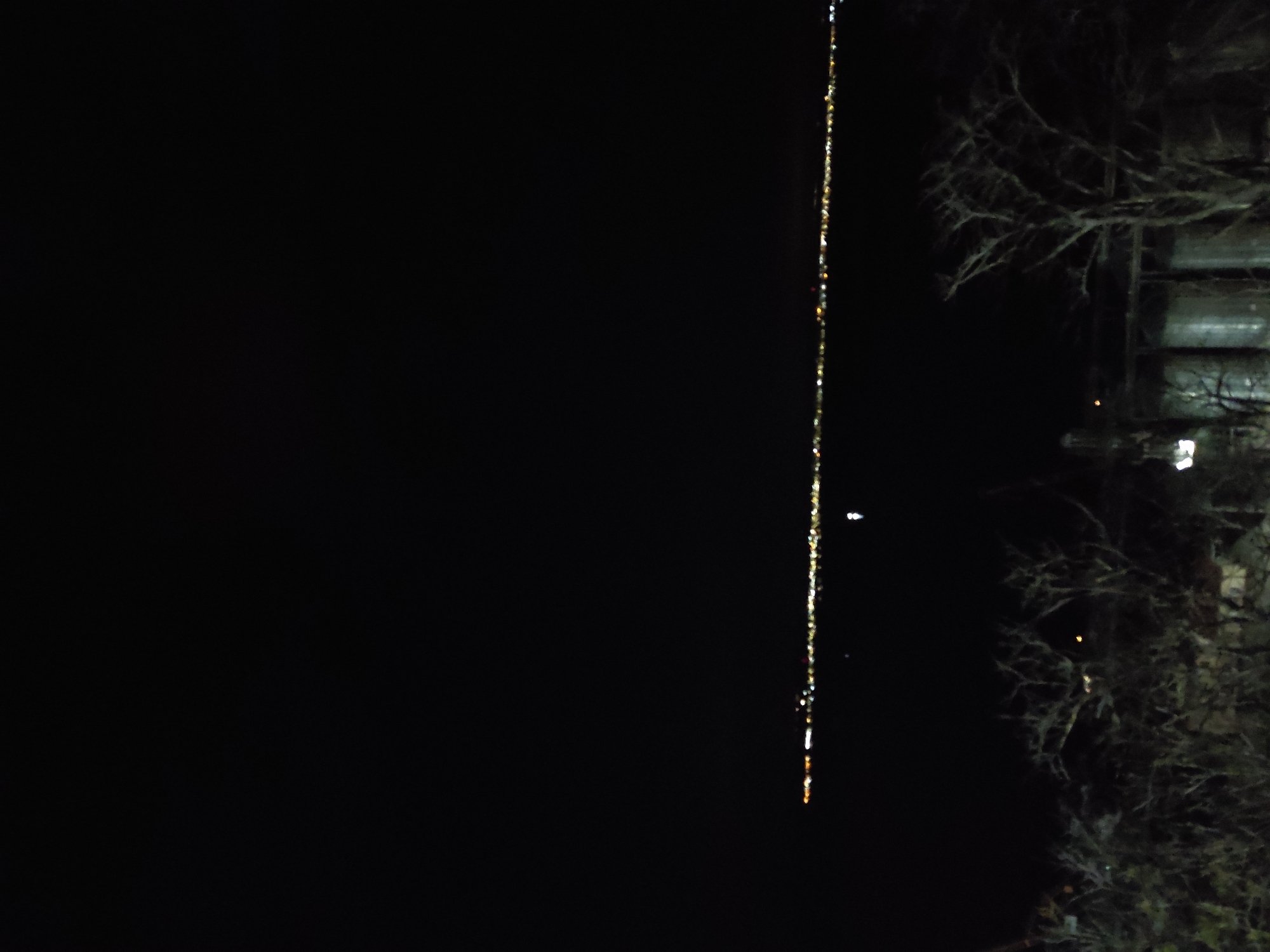 Here is a forensic description of the image.
The image appears to be a nighttime photograph taken at an outdoor location. A dark expanse covers much of the left side of the frame. A vertical line of lights bisects the frame, suggesting a reflection in water or possibly a source of lights directly. To the right, trees with bare branches are visible. There is also a structure or building, possibly with containers, that is present. The scene is dimly lit.