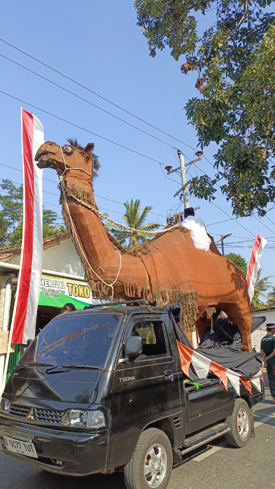 The image, captured in Krajan Lor, Indonesia, depicts a festive outdoor scene dominated by a large, tan-colored, camel-shaped float atop a black pickup truck. The float, constructed from rough material resembling burlap and adorned with fringed trim, has a rider dressed in a white shirt and a black cap seated on its back. A red and white Indonesian flag is prominently displayed to the left of the float, and a smaller red and white striped flag is attached to the rear of the truck.
The truck, identified as a Mitsubishi T120ss by its badging, is parked on a paved road. In the background, a building with a green awning displaying the word "TOKO" (shop) and text indicating "PERALATAN LISTRIK & ELEKTRONIK" (Electrical & Electronic Equipment) is visible. Palm trees and leafy trees are interspersed with utility poles and power lines against a clear blue sky. Two individuals, one in a green shirt and the other wearing a black and white patterned garment, are partially visible on the right side of the image, observing the scene. The overall impression is of a local procession or celebration.