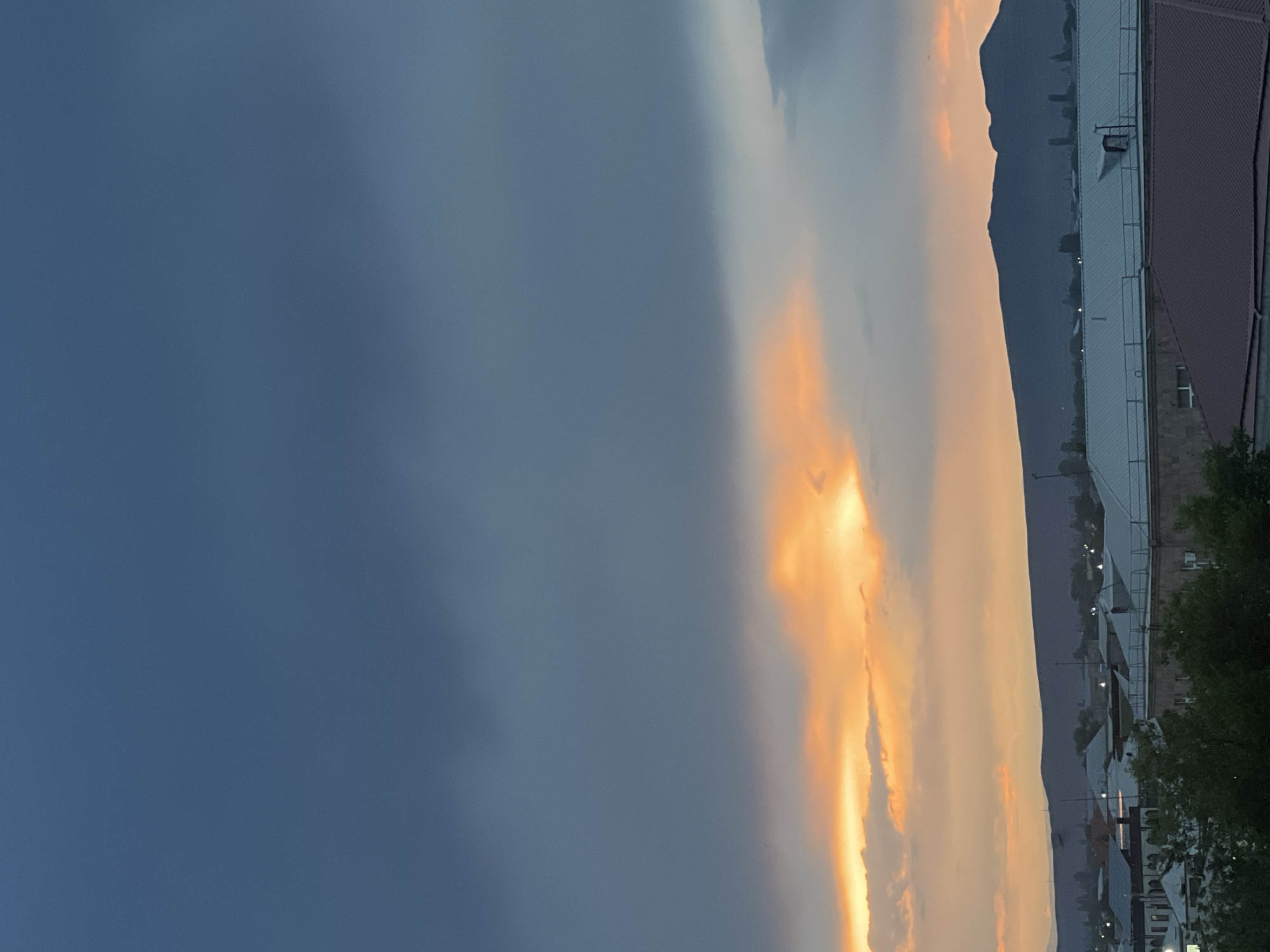 The image depicts a static scene at sunset over the city of Vagharshapat, Armenia. The upper portion of the sky is dominated by dark, desaturated grey-blue clouds. Below these, a horizontal band of vibrant orange and yellow light from the setting sun illuminates lower-lying clouds, creating a distinct contrast with the darker sky above. In the distant background, a dark, silhouetted mountain range spans the horizon, obscuring any features beyond it. The midground and foreground reveal an urban landscape consisting of numerous buildings with various roof types, including flat grey and pitched reddish-brown structures. Green tree foliage is visible in the lower left and central foreground, interspersed among the buildings. No individuals, vehicles, or discernible activities are present within the frame.
