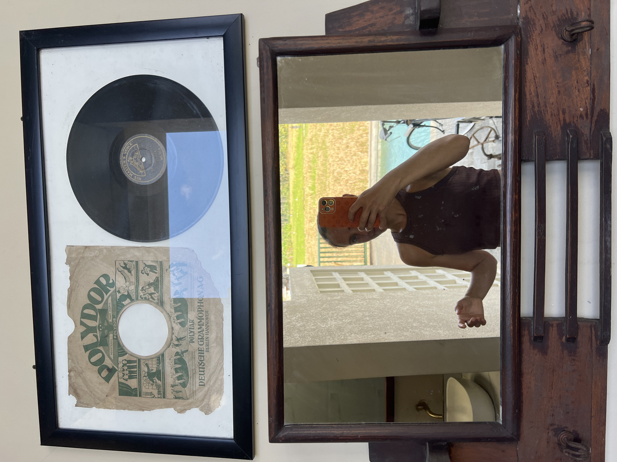 The image shows a wall with two decorative items. On the left, there's a framed vintage record, including the vinyl and its paper sleeve. The sleeve has the brand "Polydor" prominently displayed, along with some decorative illustrations and the text "Deutsche Grammophonag Berlin-Hannover."
To the right is an ornate, wooden-framed mirror. Reflected in the mirror is a person holding an orange phone, taking the picture. In the background, visible through a window, are bicycles, a tan-colored building, and greenery.
Based on the environment reflected in the mirror, this image appears to be taken in Ubud District, Indonesia.