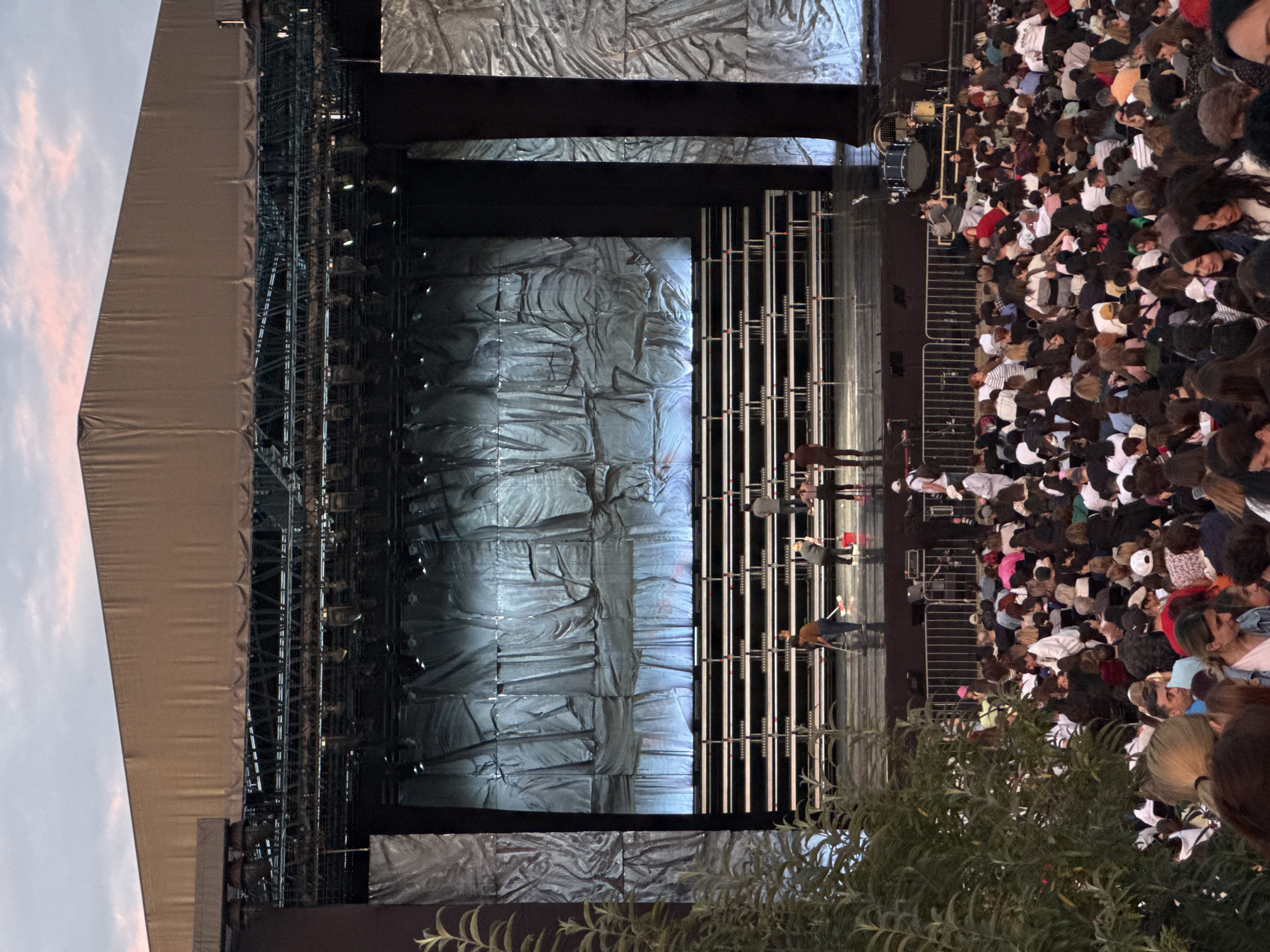 An outdoor concert or performance venue in Yerevan, Armenia, likely situated within the Cascade Complex area, is depicted at dusk. A large, semi-permanent stage structure with a beige fabric roof, supported by extensive metal trussing and lighting equipment, occupies the upper portion of the frame. The stage floor is dark and reflective, featuring multiple rows of empty tiered risers with metal frames and light-colored surfaces, presumably for a choir or orchestra. The stage's rear and side backdrops consist of large, textured grey and silver fabric panels, some illuminated by cool blue light from below.
Approximately five individuals, identified as stage crew, are on the stage floor, actively engaged in cleaning using mops and a red bucket. A drum kit is partially visible on the stage's far right. Metal crowd control barriers separate the stage from a large, densely packed audience filling the lower half of the image. The audience members are dressed in casual attire, facing the stage. Green foliage from a bush or tree partially obscures the lower-left foreground. The sky above is cloudy, exhibiting soft pink and orange hues indicative of sunset or twilight.