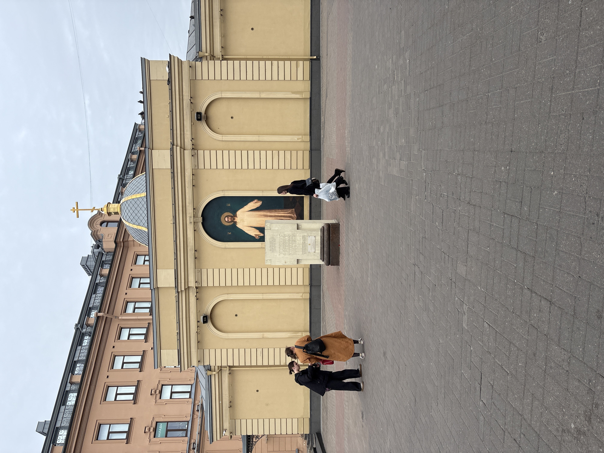 The photograph, taken during the day, shows a section of what appears to be a city square in Saint Petersburg, Russia, specifically the area around the Resurrection Church. The environment consists of the pavement made from square blocks and segments of buildings with notable architectural features.
Several people are present, including two individuals standing close to each other on the pavement, potentially engaged in a conversation or taking a photograph. Another group of two people are standing slightly farther.
A focal point is the wall of the Resurrection Church. It includes a mosaic-style rendering of a saint or religious figure, presumably Jesus Christ.
The visible section of the church features architectural details like arched windows or alcoves, a cross-topped dome and an adjoining structure with windows.