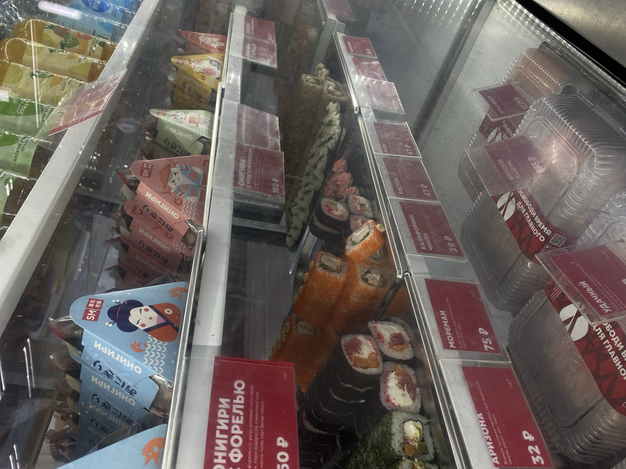 The image shows a refrigerated display case in a store, likely a grocery store or convenience store, selling sushi and other Japanese food items. The display is well-lit, and the products are arranged in an organized manner.
In the foreground, several trays of sushi rolls are visible. Some are large, uncut rolls wrapped in dark seaweed, while others are cut into individual pieces, showcasing colorful fillings. There are also smaller triangular packages of onigiri, a type of Japanese rice ball, with various designs and illustrations on the packaging.
Above the sushi, another shelf displays more onigiri packages, as well as bottles of drinks. The drinks appear to be juices or teas, with labels in Russian.
Several price tags are visible throughout the display, indicating the cost of the items in Russian rubles. The overall impression is of a modern, clean, and well-stocked food display.
It is not possible to determine the specific location within Peterhof, Russia from the image alone.